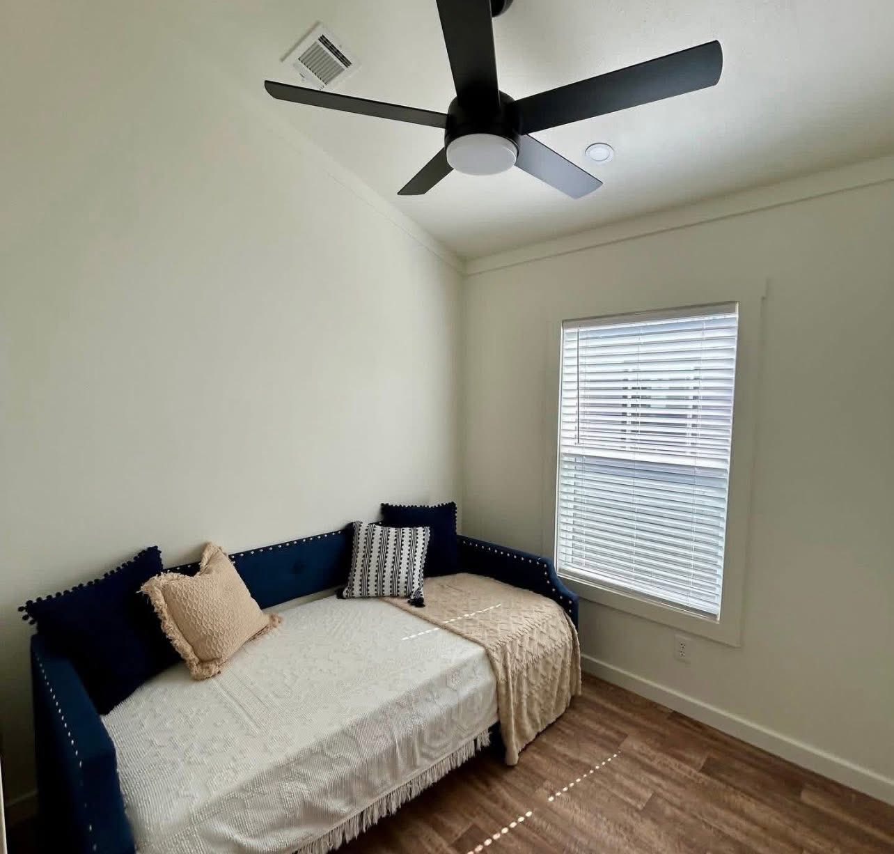 A cozy room with a minimalist vibe features a navy daybed with decorative pillows, a white window blind, a beige throw, and a modern ceiling fan.