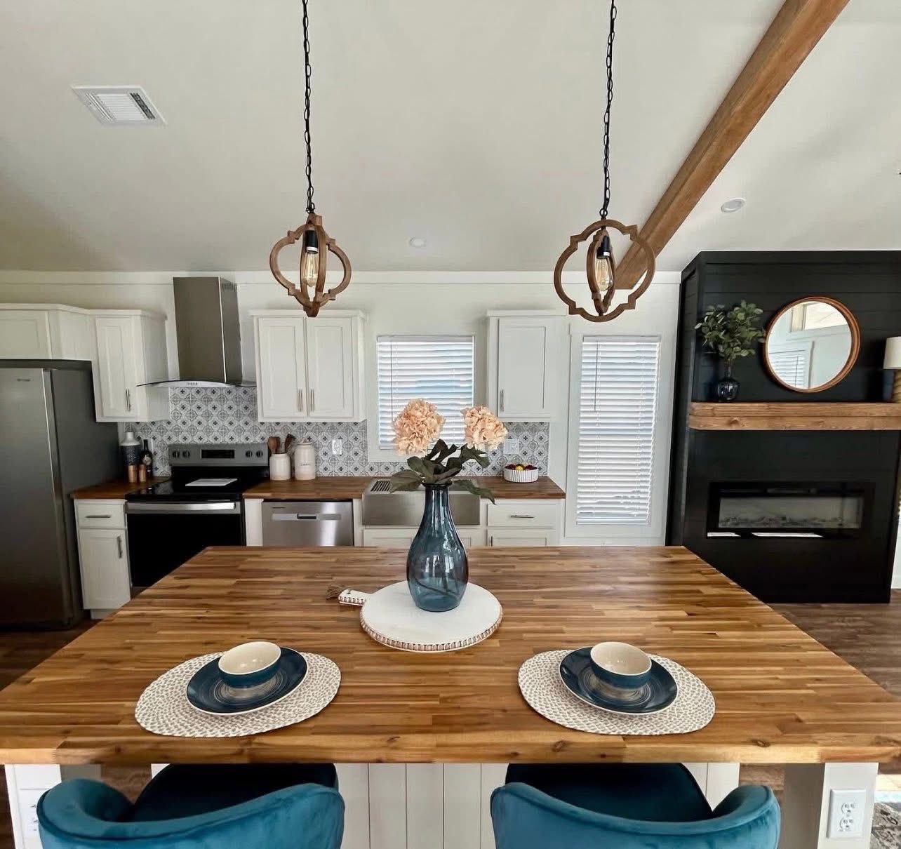 Modern kitchen with a wooden island, two teal seats, hanging lights, and white cabinets. A vase with flowers adds charm; cozy and inviting ambiance.