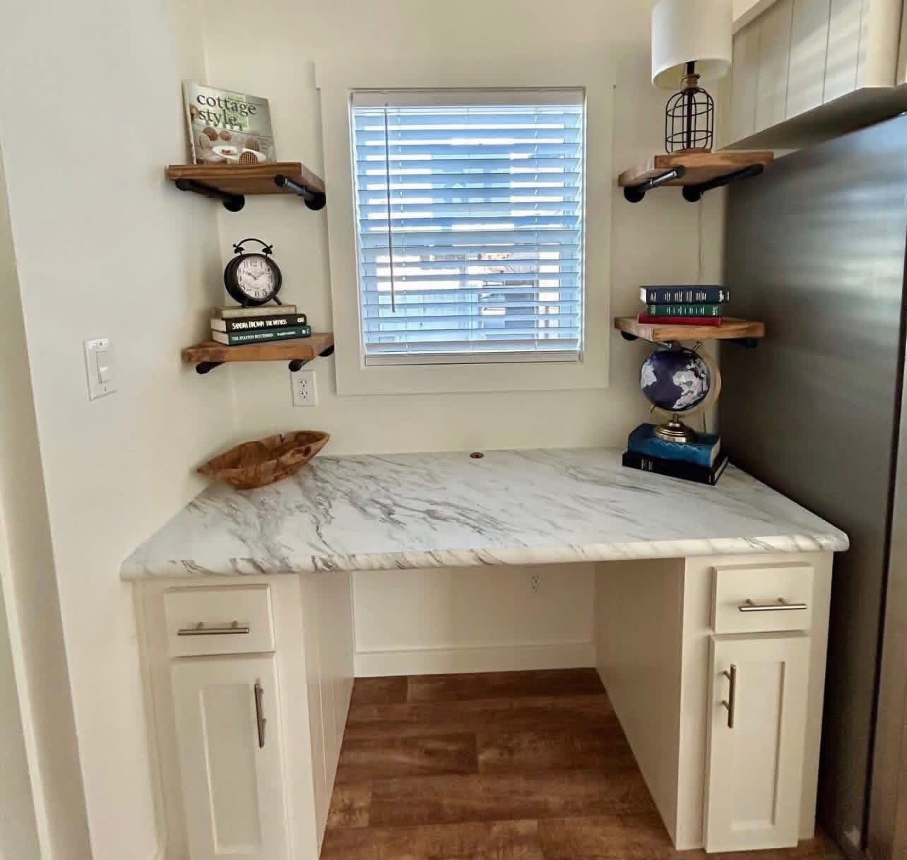 A small nook with a white marble desk, shelves with books, a globe, and a clock. A window with blinds above, creating a cozy, organized workspace.