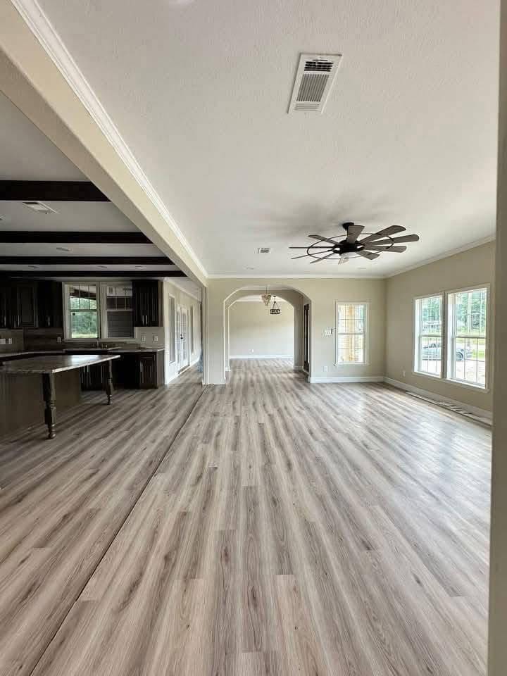 Spacious open-concept room with wooden floors and ceiling fan. Kitchen with dark cabinets and island on left, large windows on right, serene ambiance.