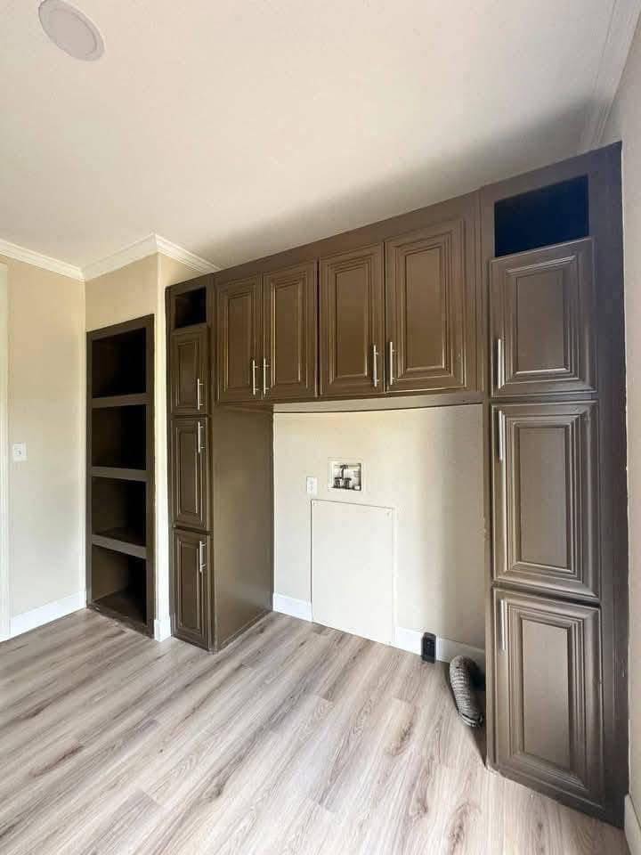 Brown wooden cabinets with silver handles line a beige wall, featuring an empty space and covered nook. Light wood floors add a modern touch.