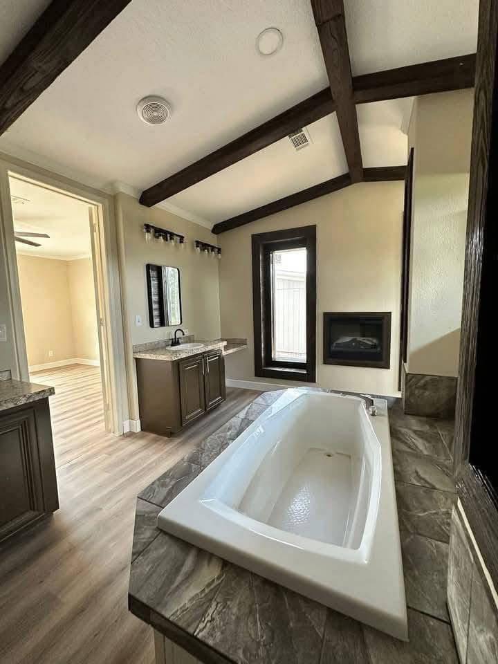 Elegant bathroom featuring a large white bathtub with stone accents, wood-beamed ceiling, double sink vanity, and a modern fireplace by a tall window.