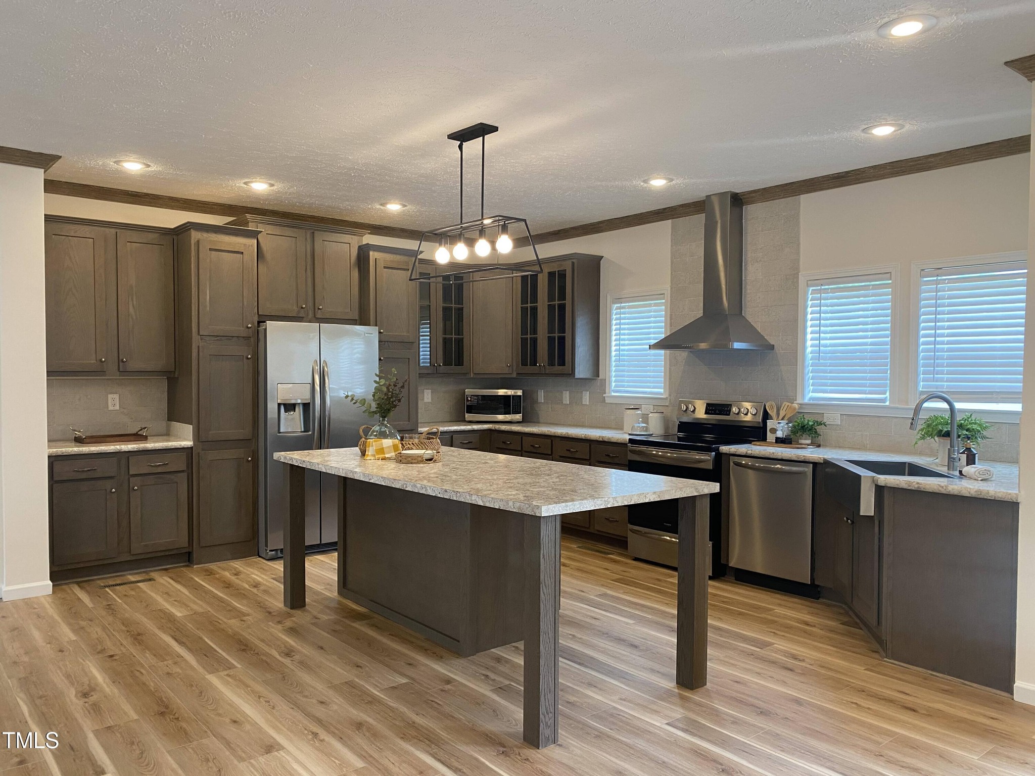Spacious kitchen with dark wood cabinets, stainless steel appliances, and a large island with pendant lighting. Light wood flooring and bright windows add warmth.