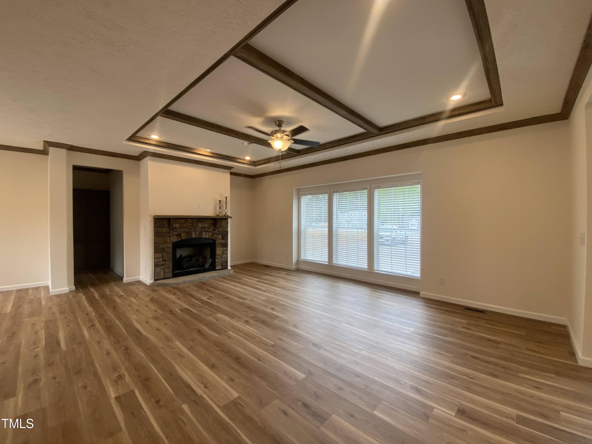 Spacious living room with wood flooring, a tray ceiling, and a ceiling fan. A stone fireplace is set against one wall, and large windows provide natural light.
