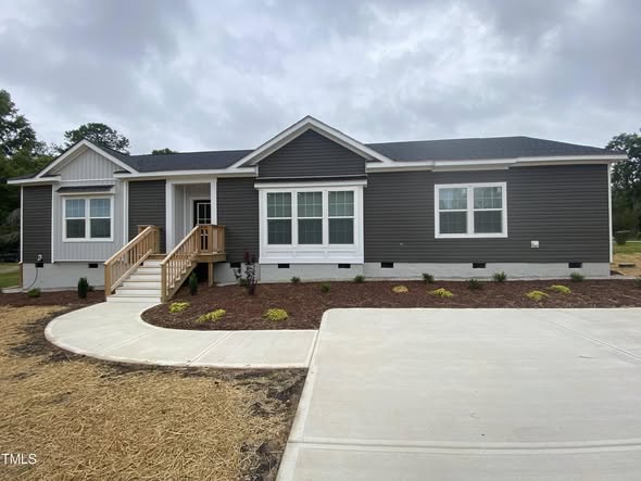 A modern, single-story gray house with white trim. Features include a small wooden porch, a neat driveway, and landscaped garden beds under a cloudy sky.