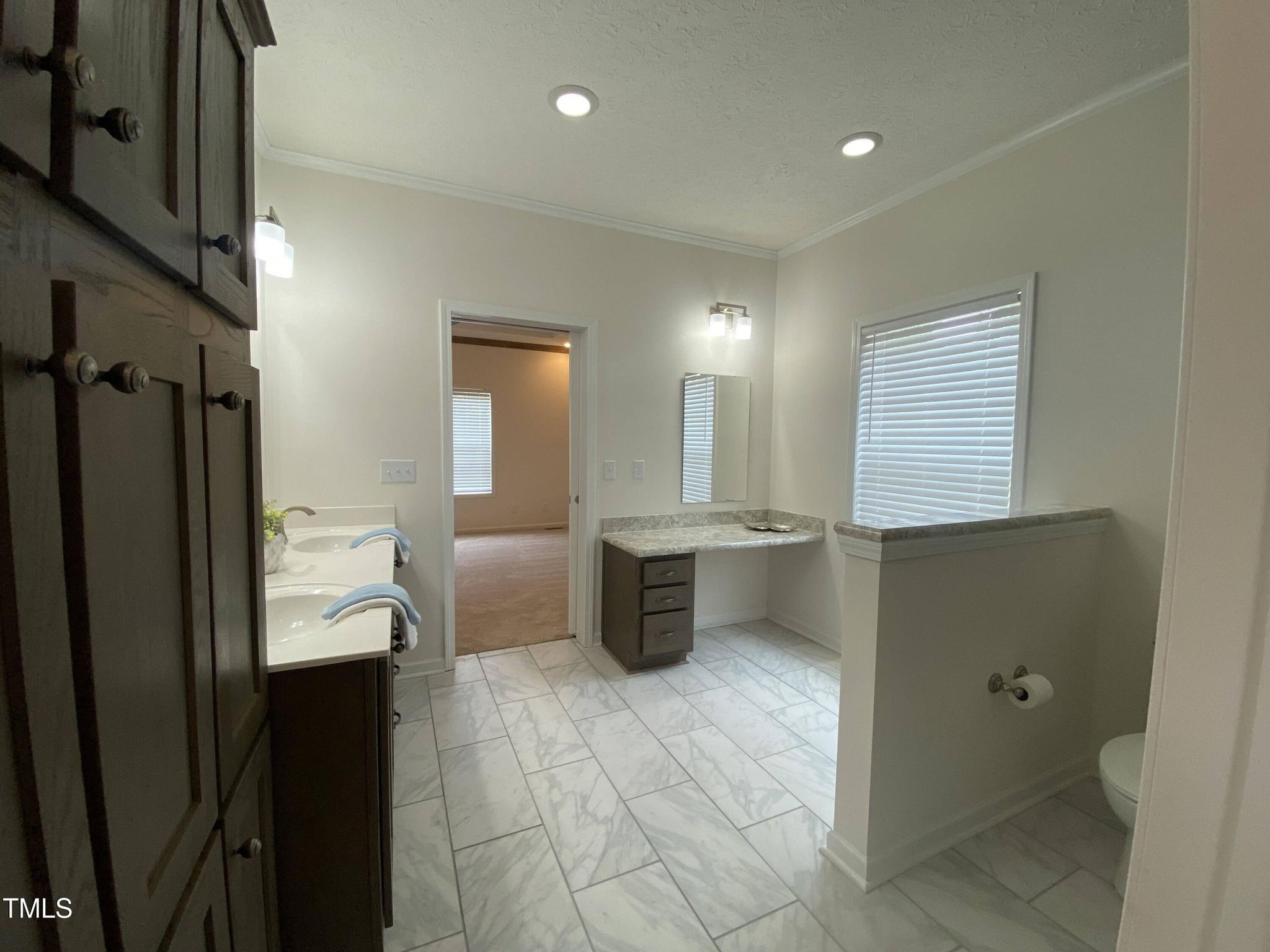 Modern bathroom with marble tile floor, dual sinks with dark wood cabinets, and a small partitioned toilet area. Bright lighting and neutral tones.