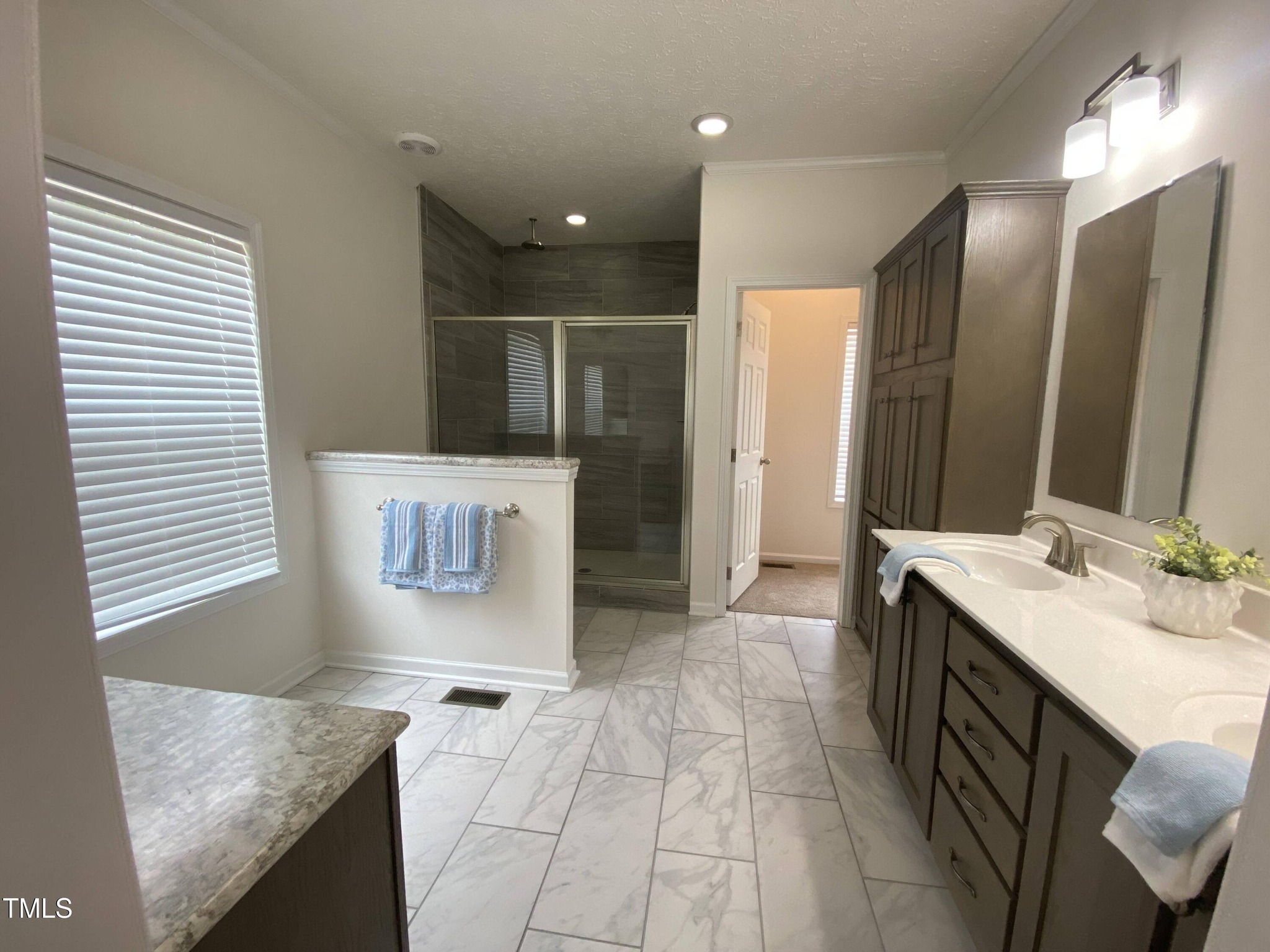 Spacious bathroom with marble tile floor, large glass shower, double vanity with brown cabinets, modern fixtures, and soft lighting; serene atmosphere.