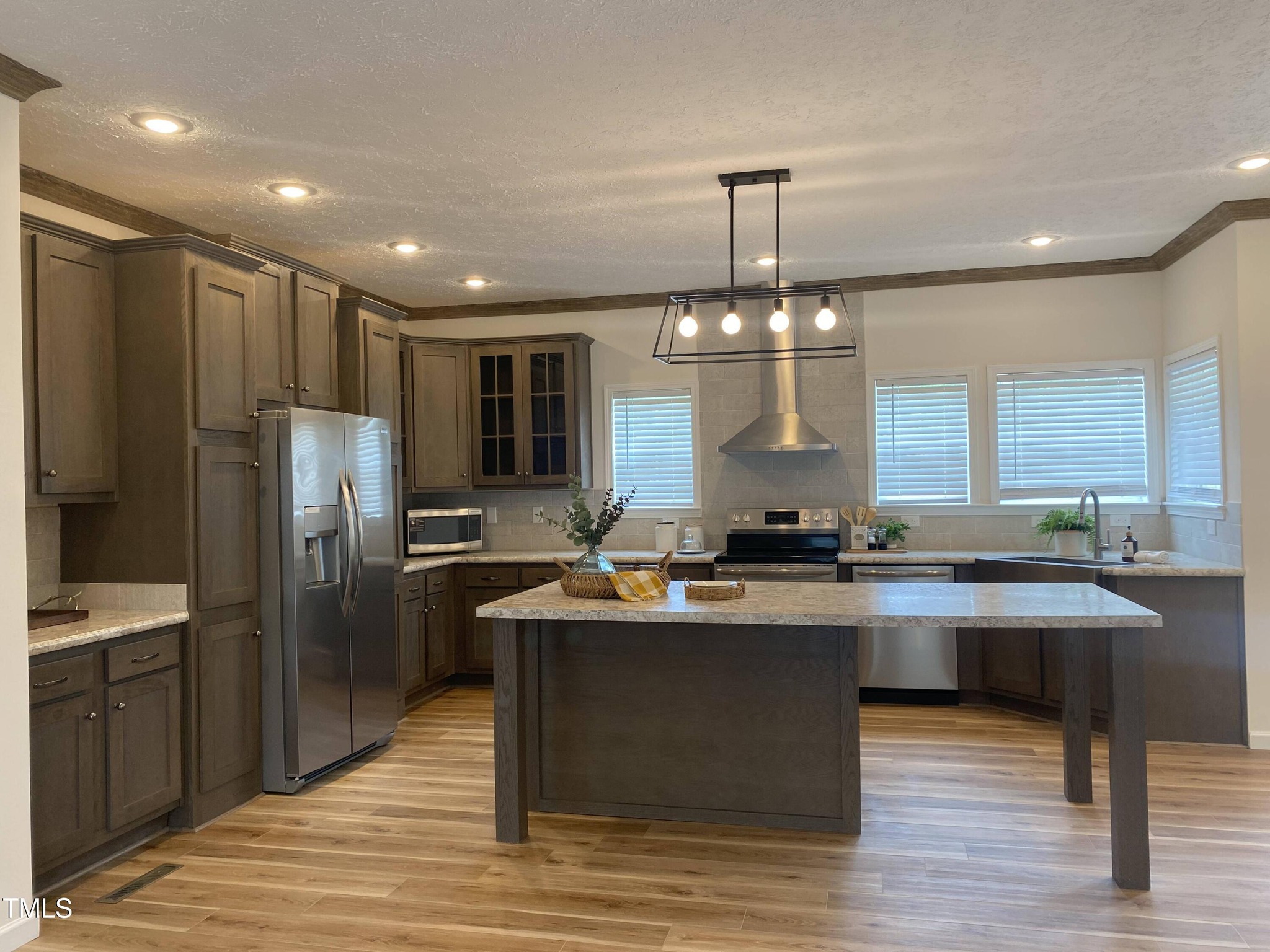 Modern kitchen with wooden cabinets, stainless steel appliances, and an island under pendant lighting. Bright ambiance with natural light and plants.