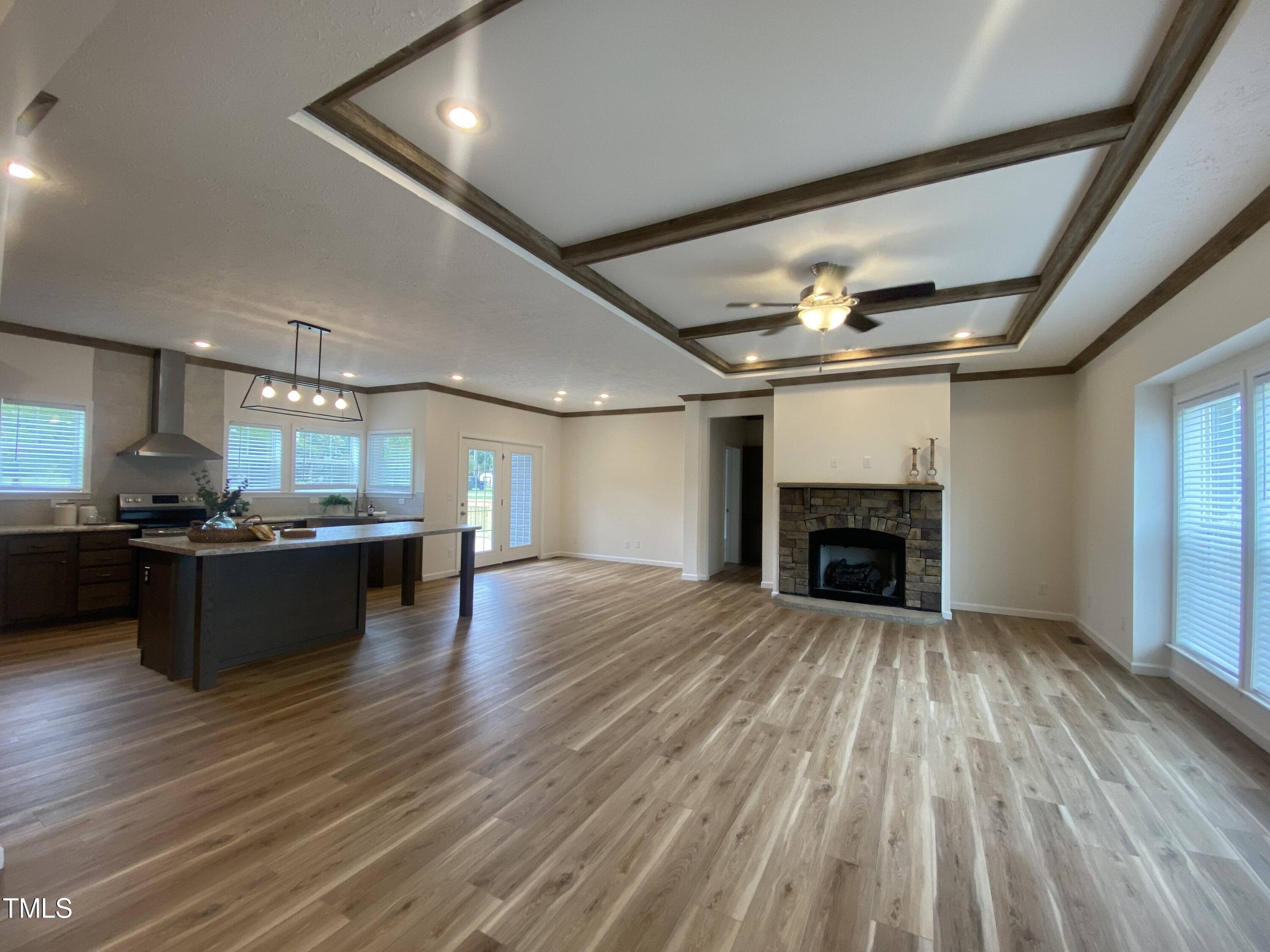 Spacious open living area with wooden floors, coffered ceiling, and a modern kitchen. Stone fireplace adds warmth. Large windows provide natural light.