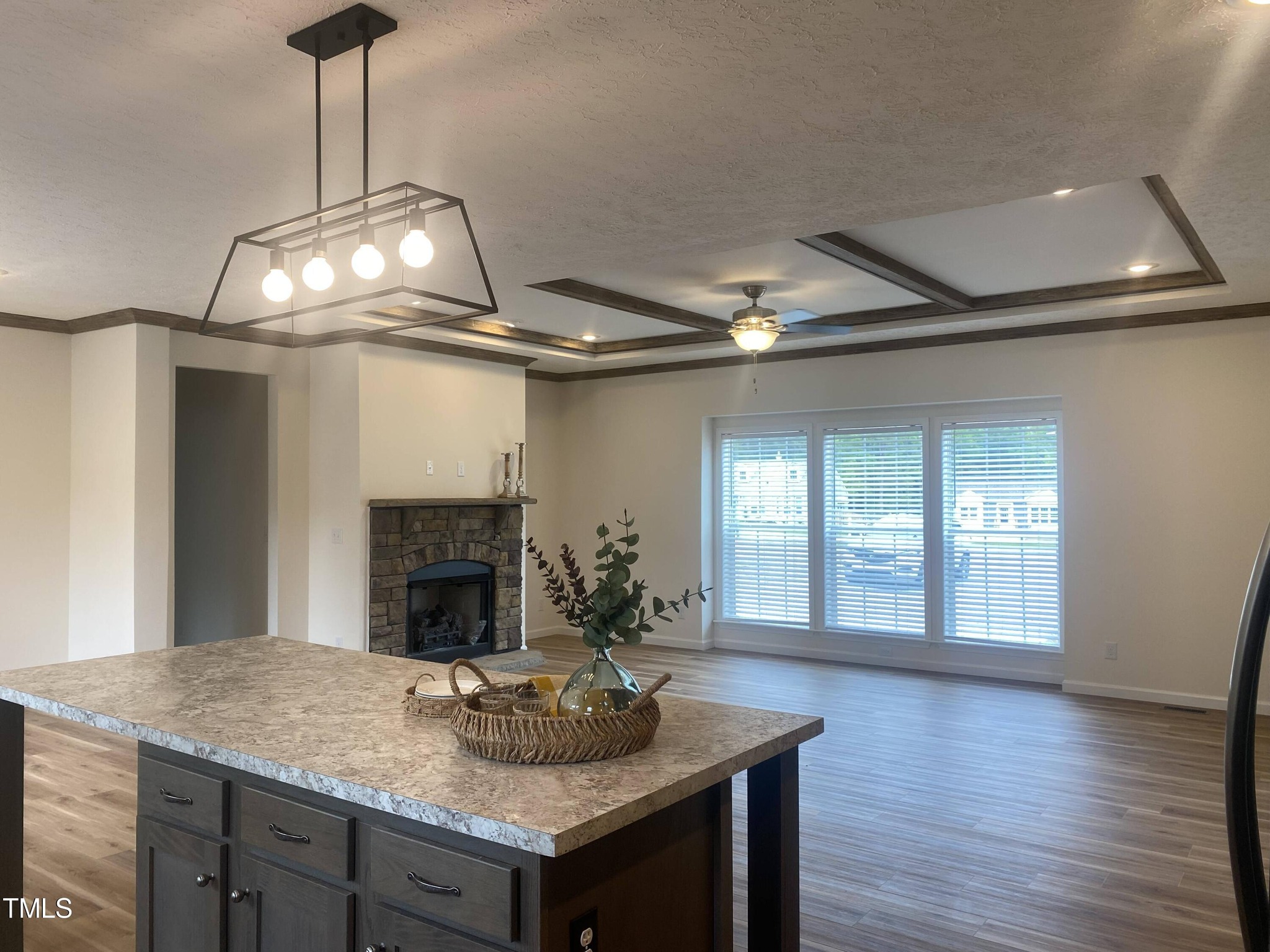 Spacious living area with light wood flooring, a stone fireplace, and large windows. Central island with decorative items, under a modern light fixture.