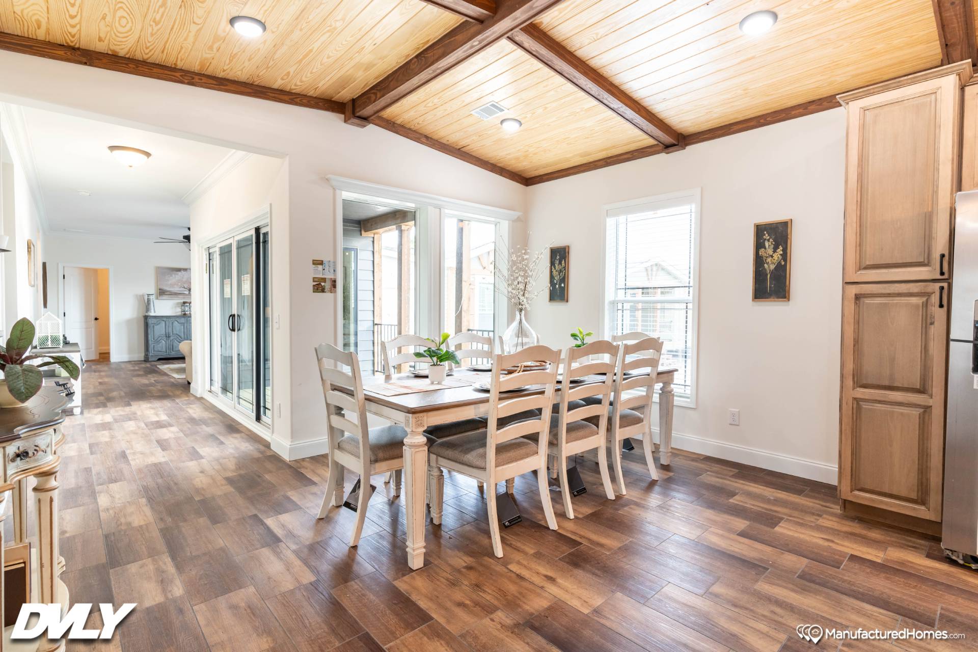 Bright dining area with a wooden table and six chairs on wooden flooring. Ceiling has wood panels and beams. Natural light streams through large windows.