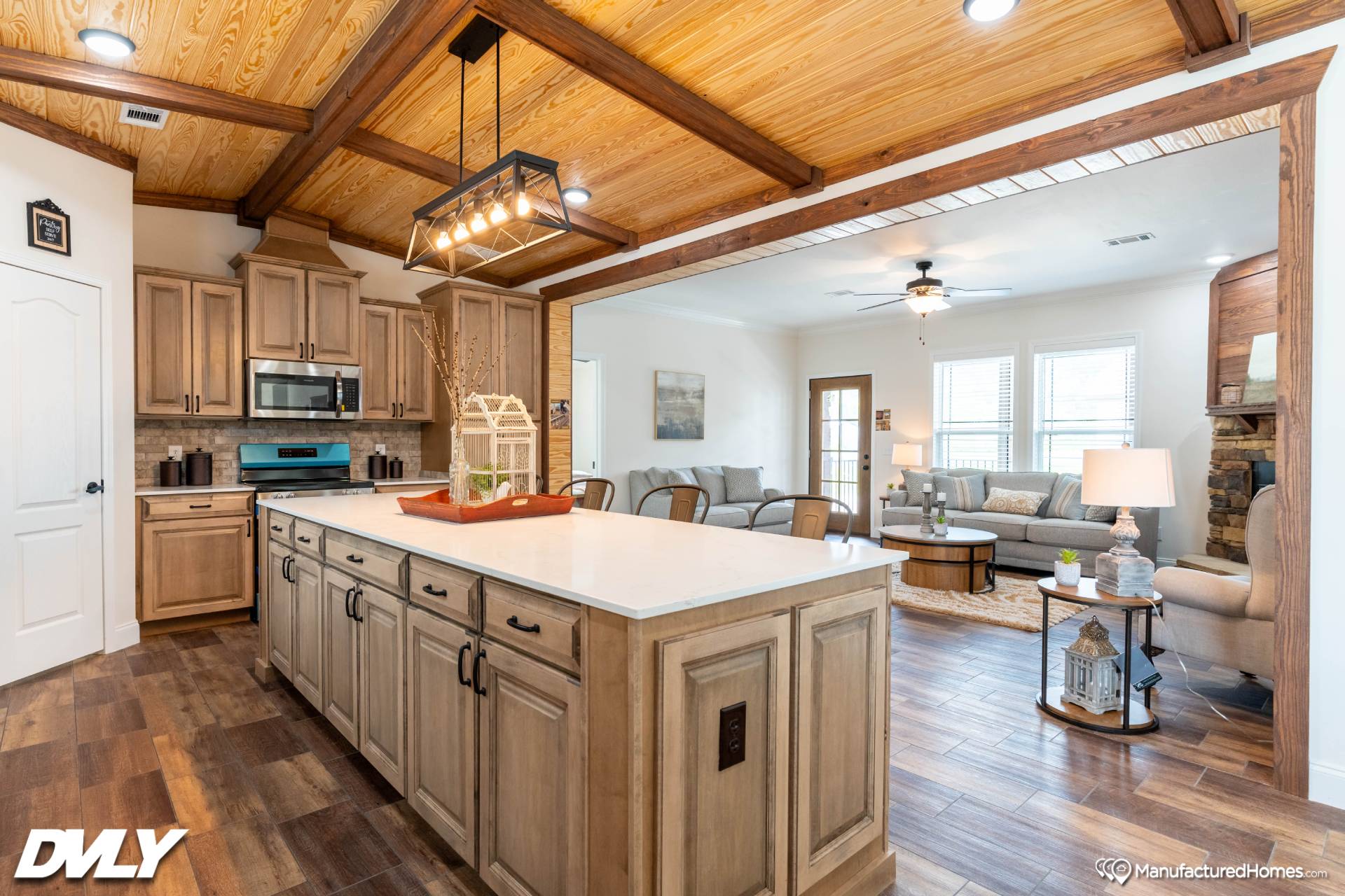 Warm, modern kitchen with wooden cabinets, large white island, and rustic lighting. Opens to a cozy living room with neutral tones and a ceiling fan.