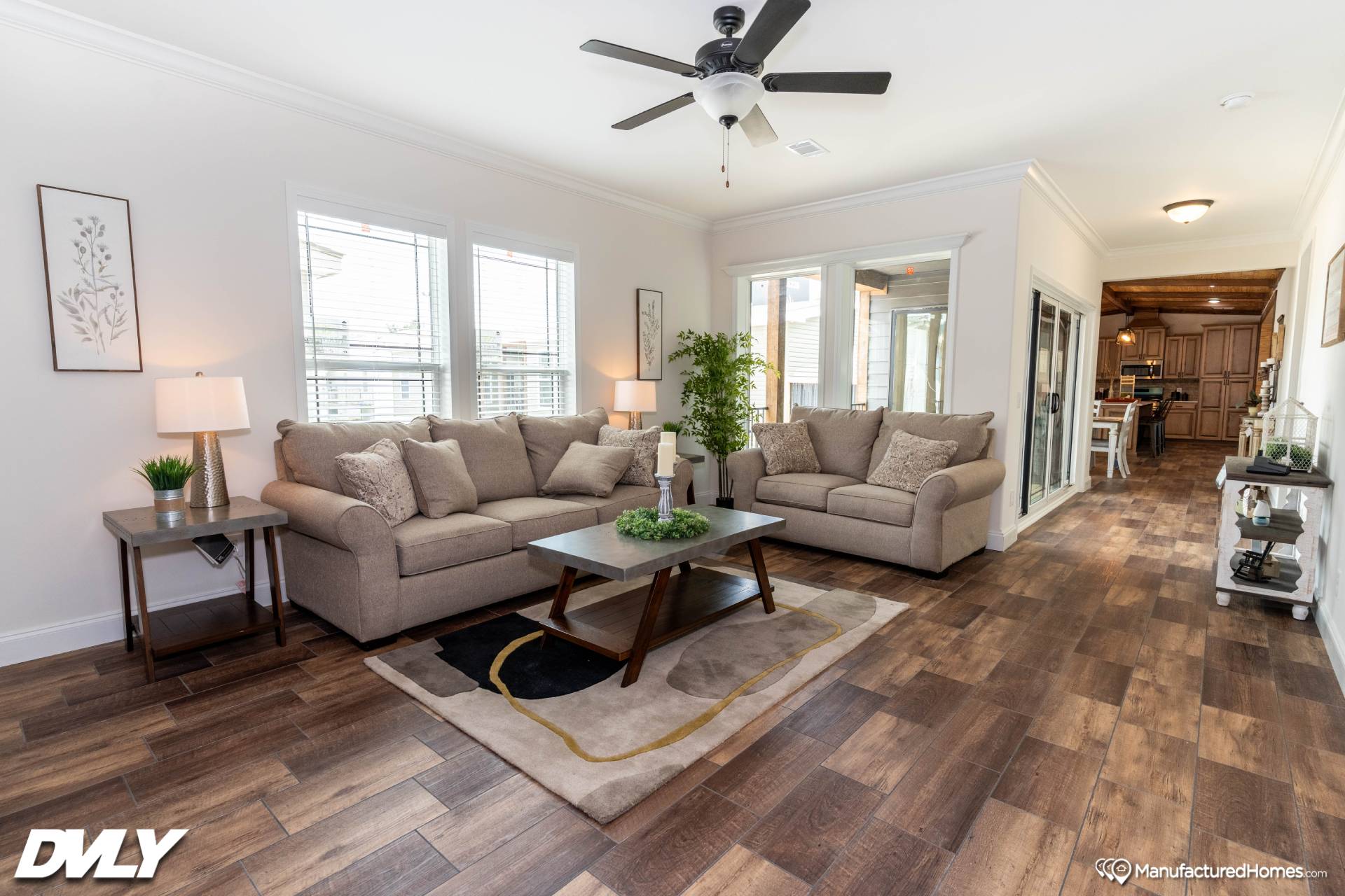 A spacious living room with two beige sofas, a gray coffee table, and indoor plants on wooden flooring. Large windows allow natural light, and a ceiling fan is centered above. The room has a cozy, modern feel.