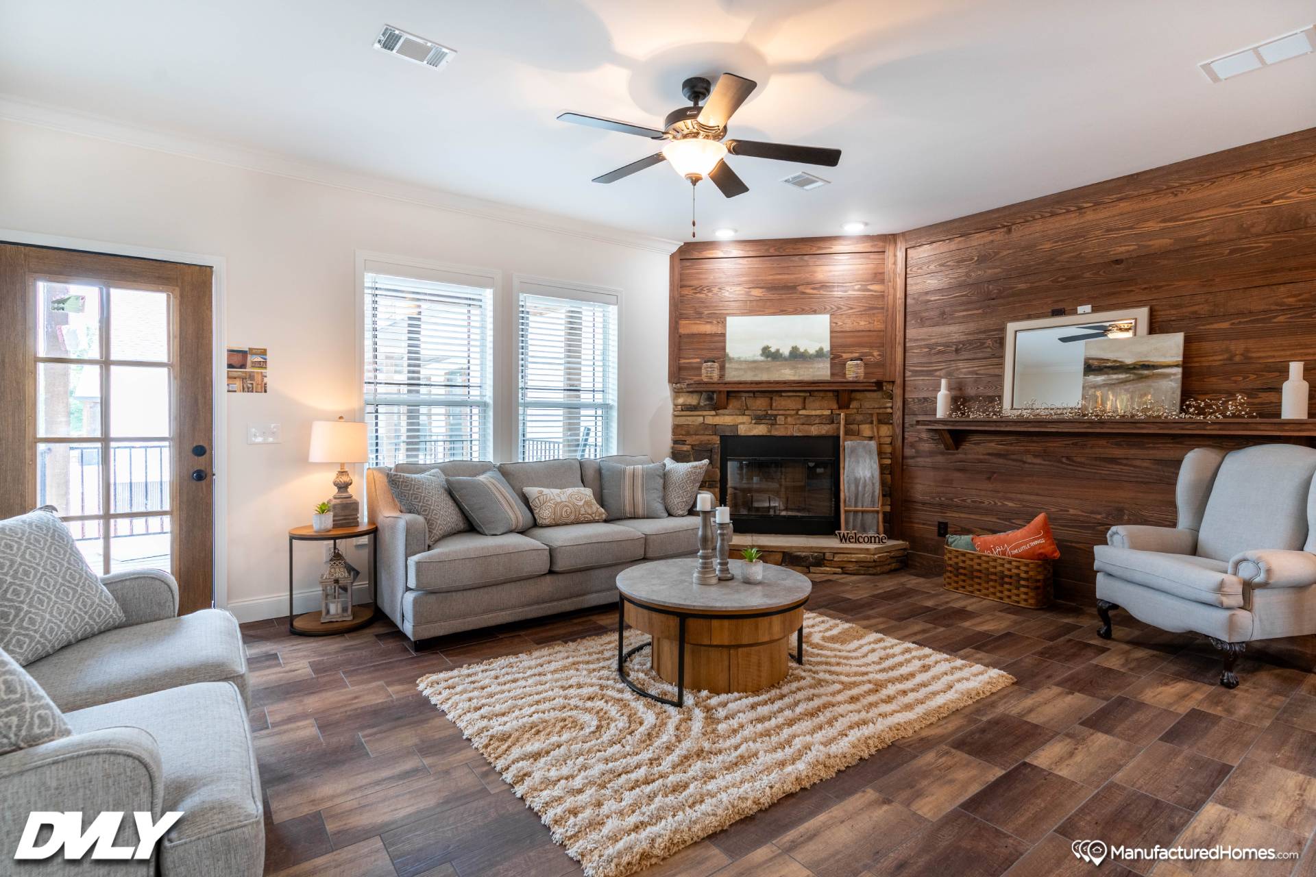 Cozy living room with gray sofas, a light rug, and a round wooden coffee table. Wood-paneled wall with fireplace and decor, ceiling fan overhead.