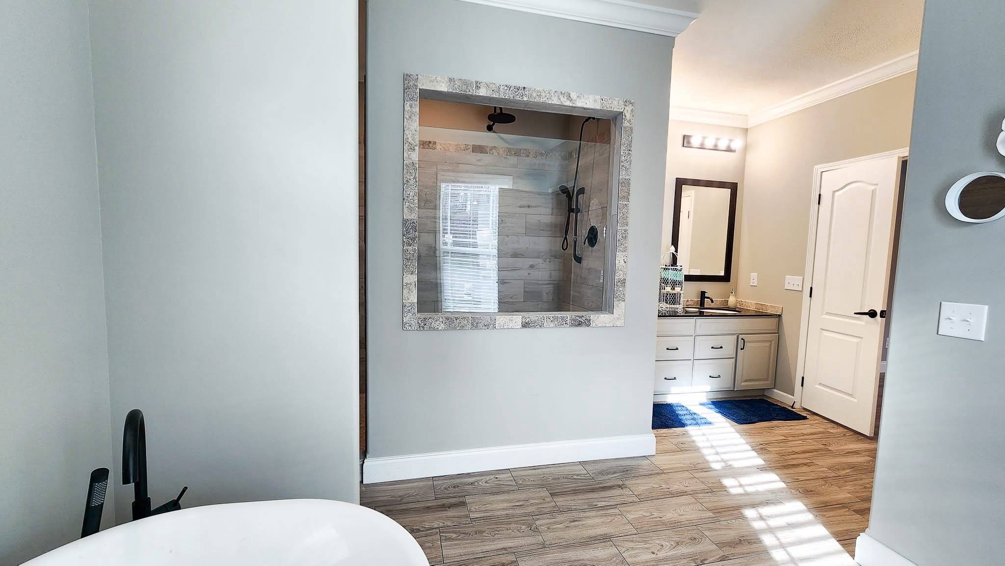 Modern bathroom with a freestanding tub, wood-look flooring, and a spacious shower with a stone frame. Vanity with mirror on the right, warm lighting.