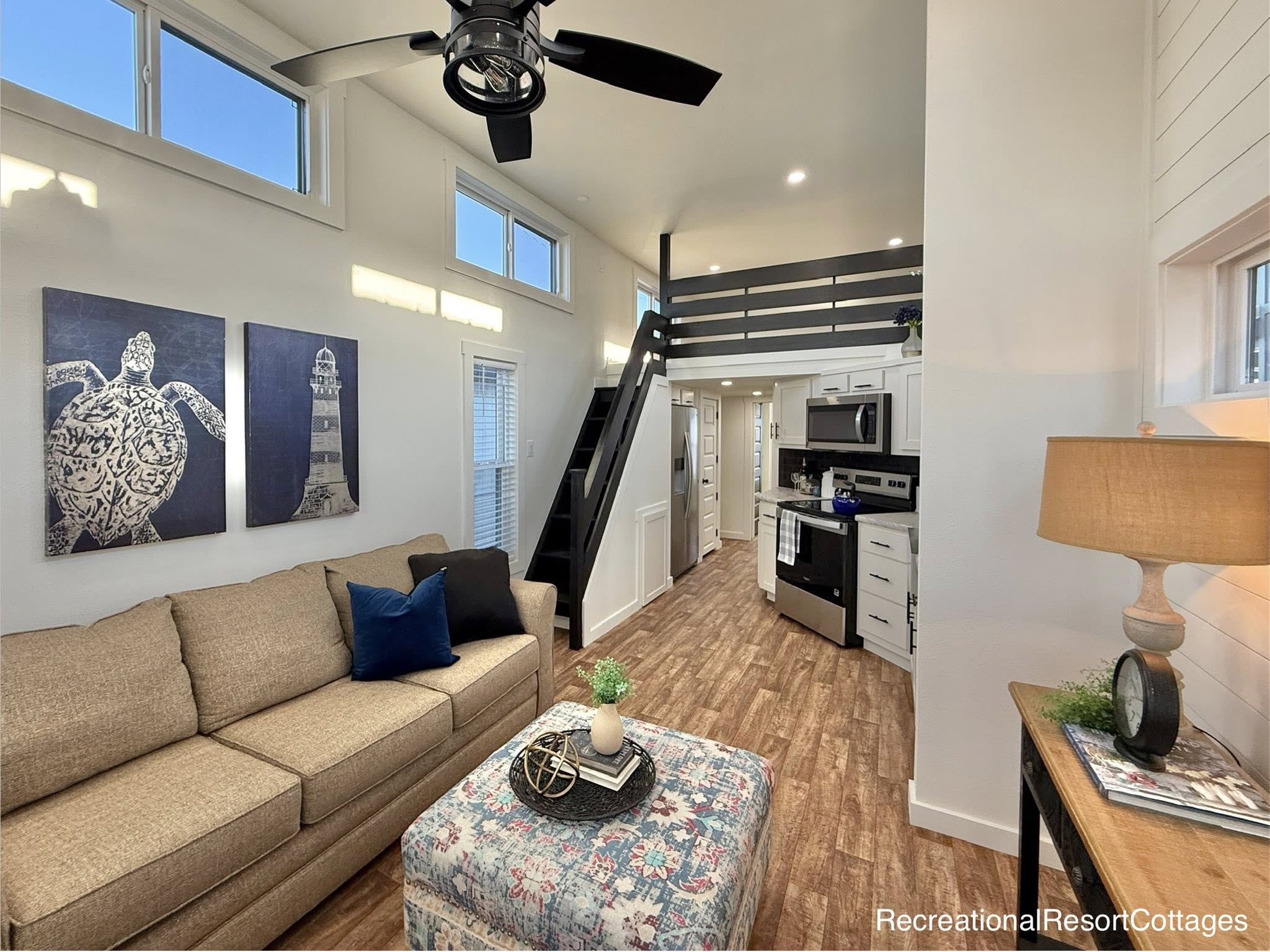 Cozy tiny home interior with a beige sofa, blue cushion, and turtle artwork. Loft with black railing, modern kitchen, and wood flooring under a ceiling fan.