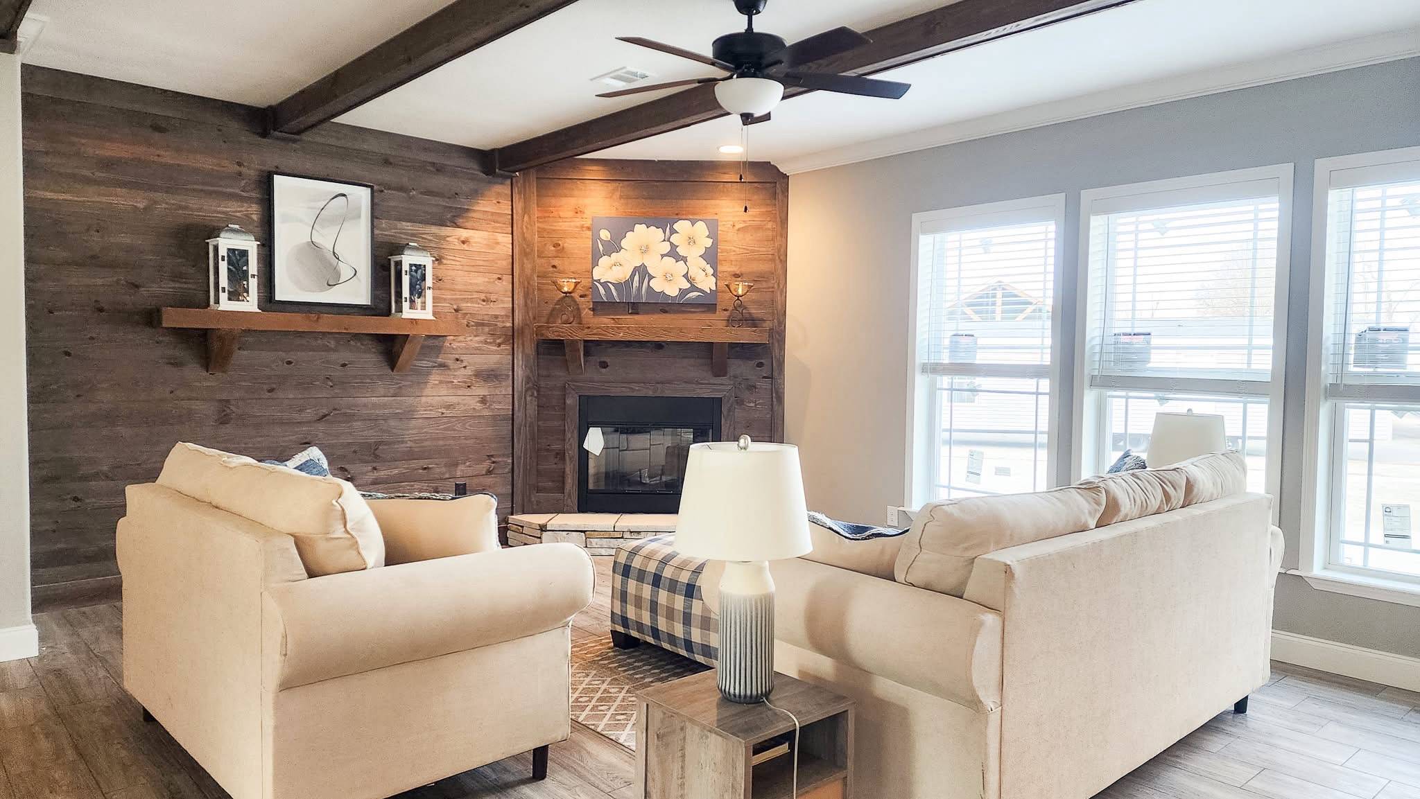 A cozy living room with a beige sofa set facing a wooden mantel fireplace. Large windows brighten the space, and floral art adds a warm touch.