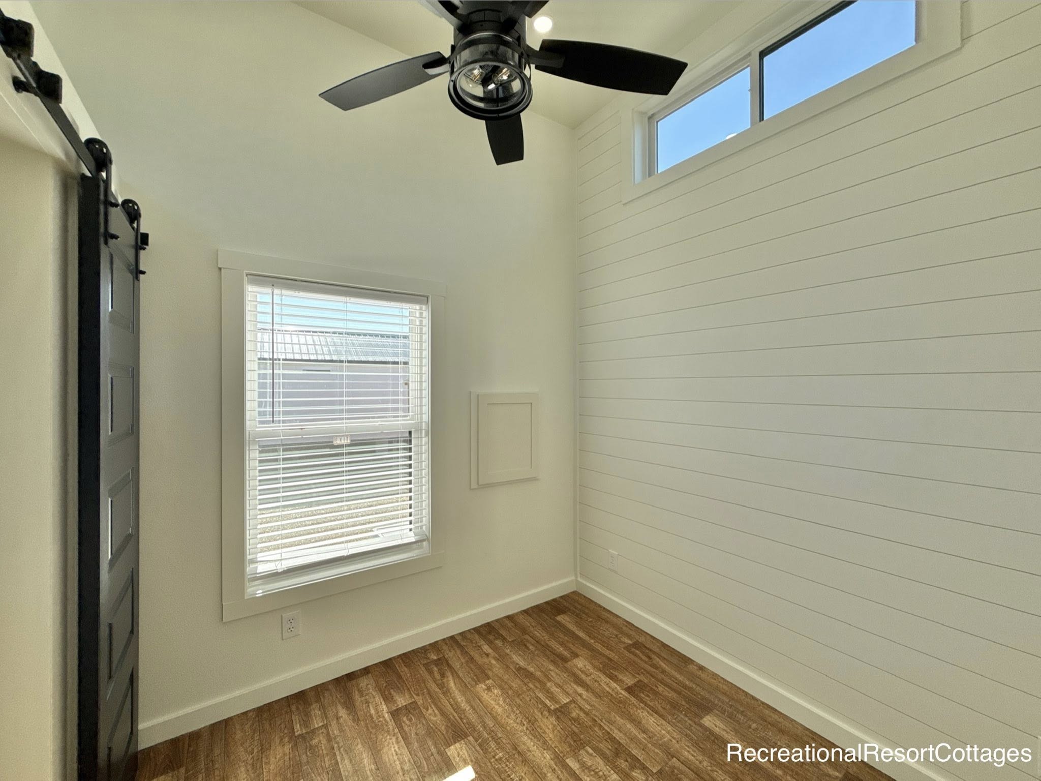 Bright room with white shiplap walls and wood flooring. A black ceiling fan hangs above, with natural light streaming through a window with blinds.