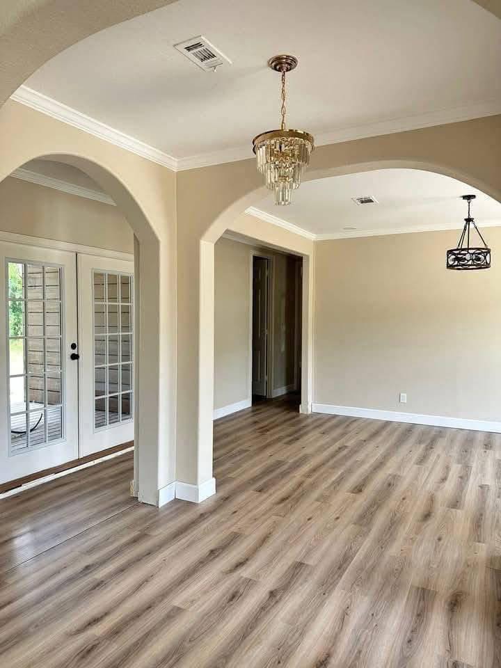 Spacious, light-filled room with light wood flooring, beige walls, and a chandelier. French doors on the left open to a patio, creating an inviting atmosphere.