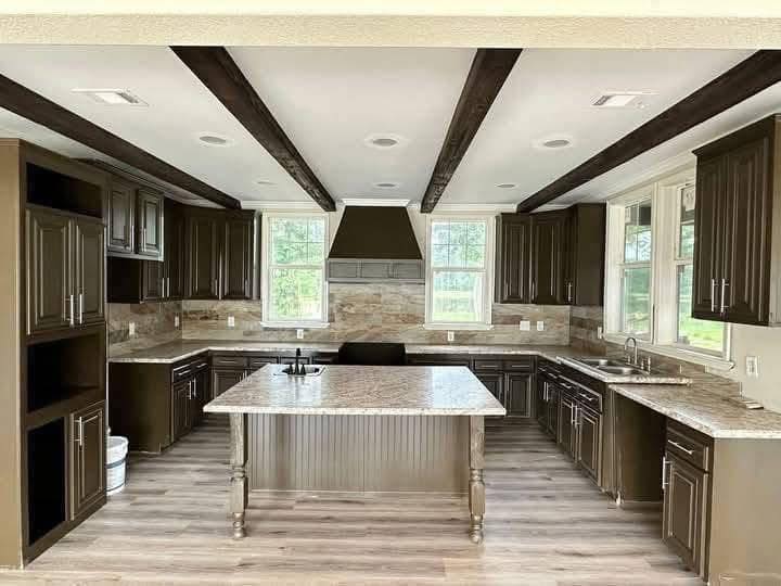 Spacious kitchen with dark wood cabinets, marble countertops, central island, and large windows. Exposed beams and light wooden floor add warmth.