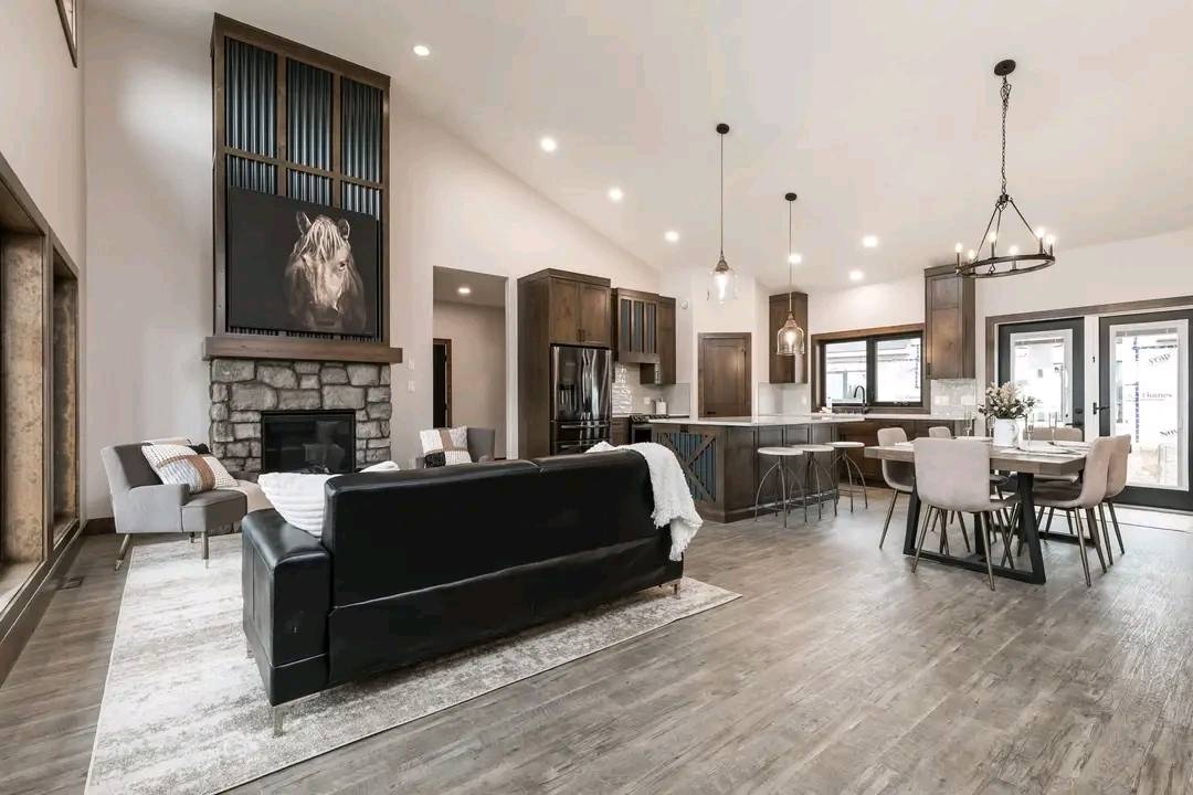 Spacious open-concept living room and kitchen with rustic decor. Features a black sofa, stone fireplace, dining table, and high ceilings. Cozy ambiance.
