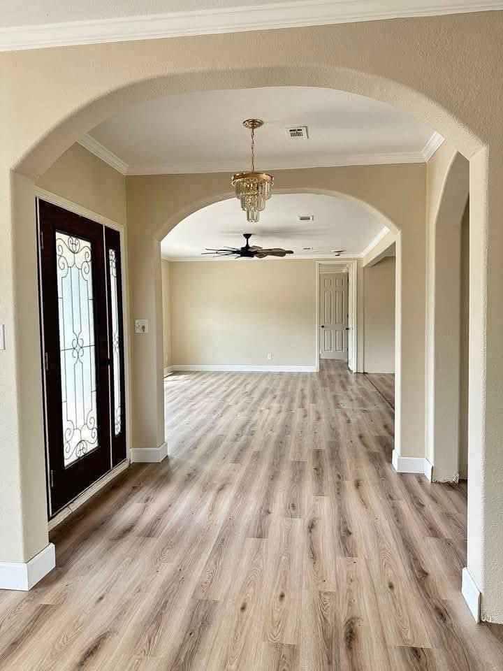 Spacious hallway with light wood floors, cream walls, and arched doorways. A chandelier hangs above, leading to a room with a ceiling fan.