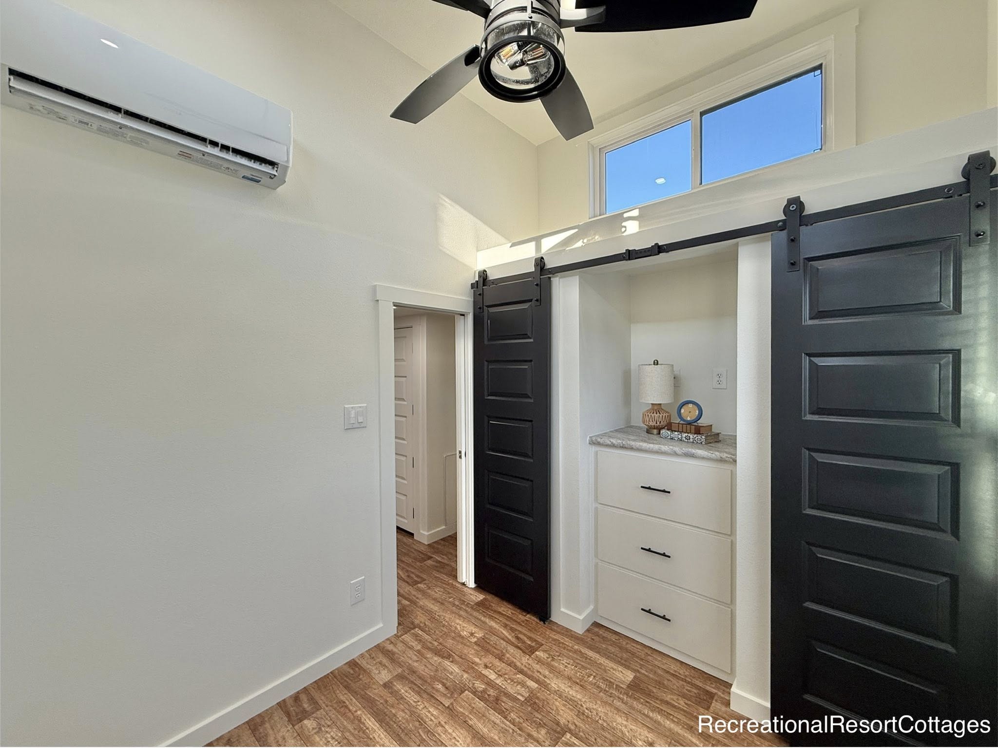 Modern room with a ceiling fan and air conditioning unit. Features sleek black sliding barn doors, white drawers with a lamp, and sunlight streaming in.