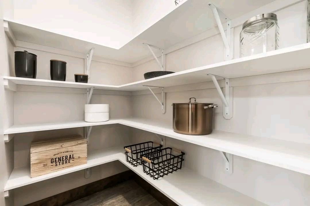 A neatly organized pantry with white shelves holds jars, canisters, and baskets. A wooden box labeled "General Store" adds a rustic touch. Clean and orderly.