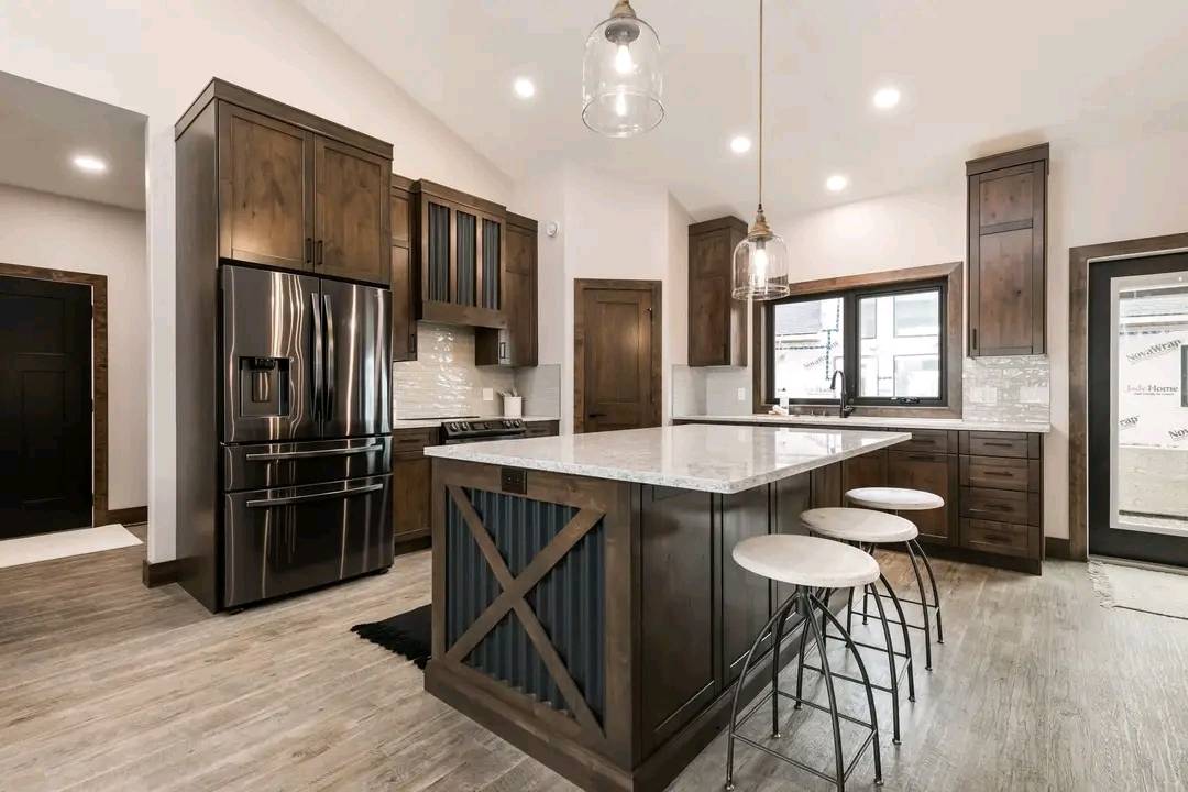 Modern kitchen with dark wood cabinets, large island with white countertop, and three stools. Stainless steel fridge and pendant lighting create a warm, inviting ambiance.