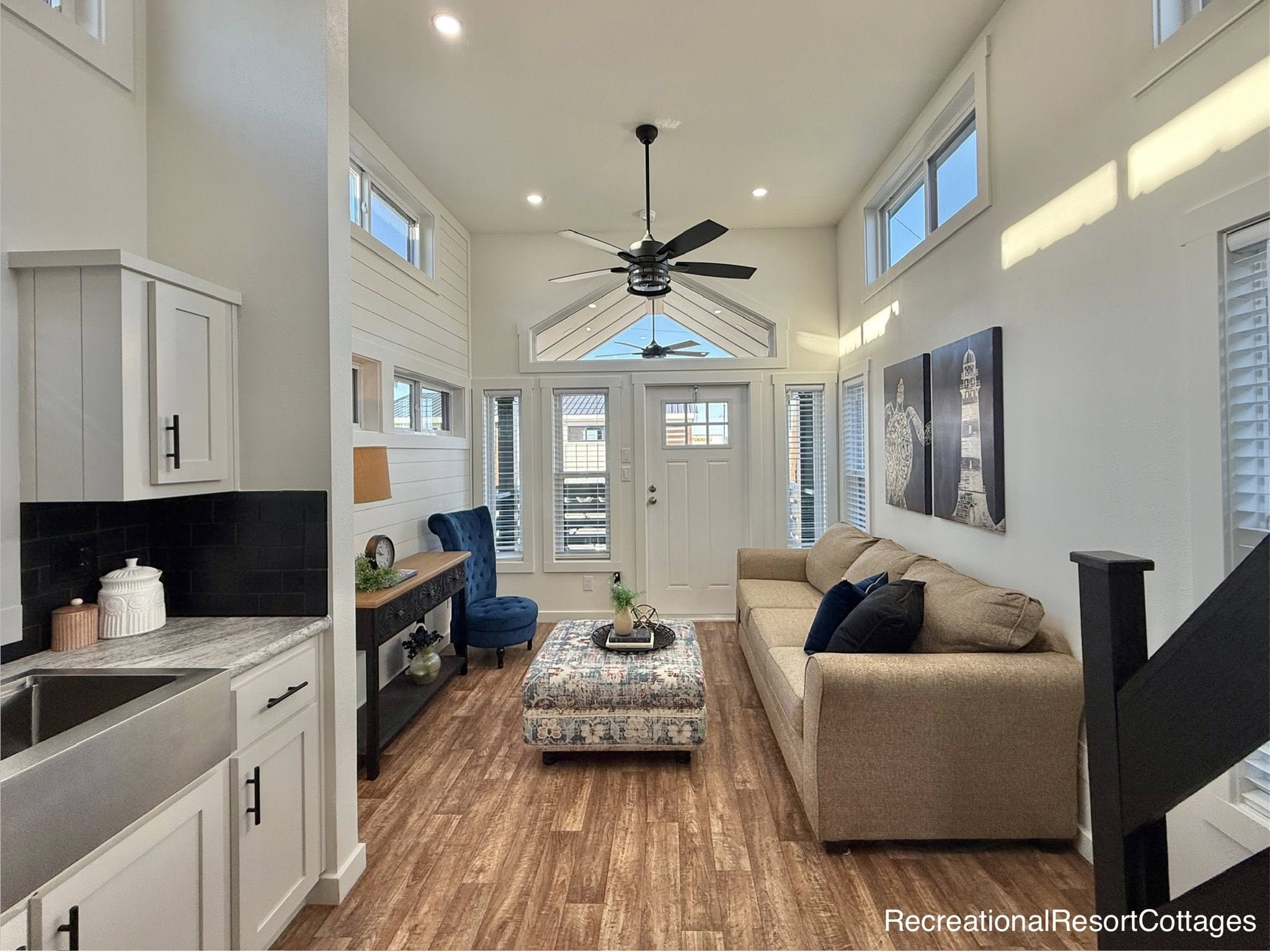 Stylish living room with high ceilings, a beige sofa, colorful ottoman, and blue chair. Bright natural light from tall windows, modern decor.