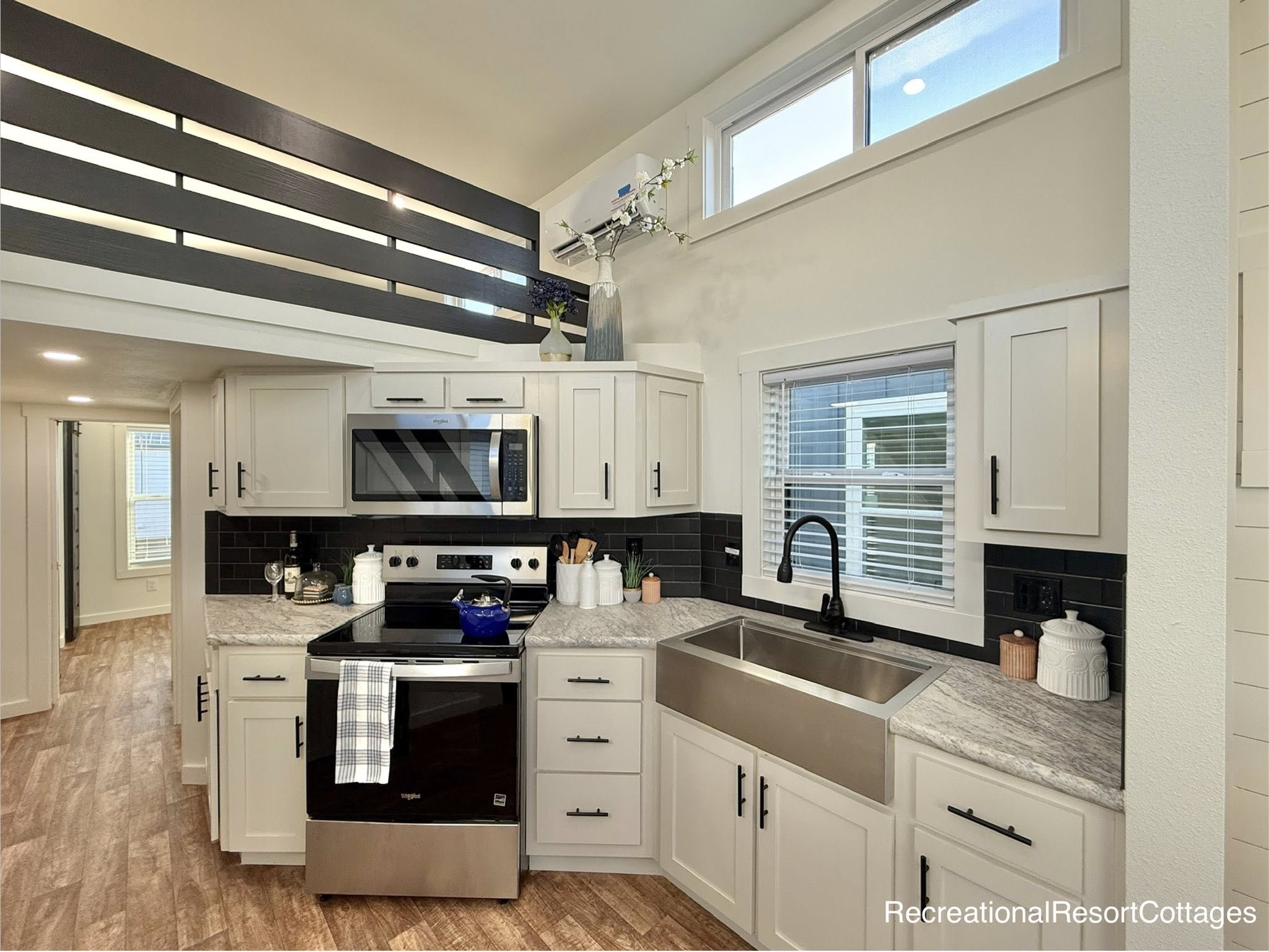 Modern kitchen with white cabinets, stainless steel appliances, and black backsplash. A large sink is under a window, with wooden floors and loft railing above. Cozy and contemporary vibe.