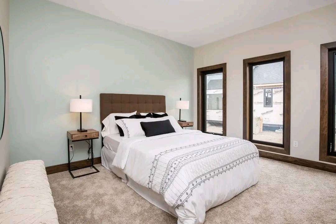 Cozy bedroom with a soft neutral palette, featuring a neatly made bed with white and black accents, dual bedside lamps, and large windows allowing natural light.