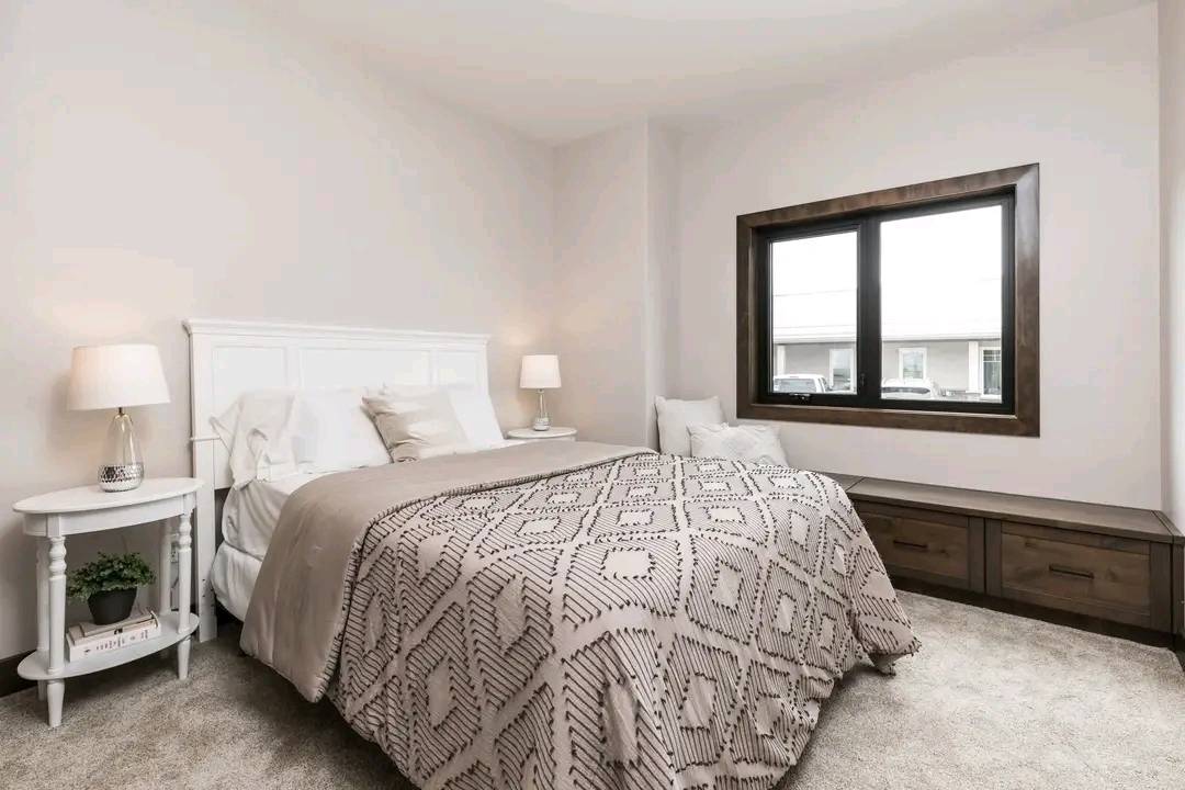 A cozy bedroom with a white headboard, patterned beige bedding, and two bedside lamps on white tables. A large window and a wooden bench add warmth.