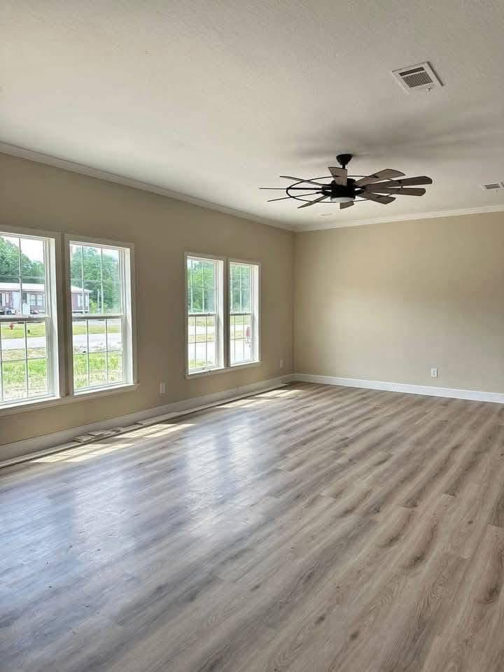 Spacious, empty room with light wood flooring, beige walls, three large windows, and a modern ceiling fan. Sunlight creates a warm, inviting glow.