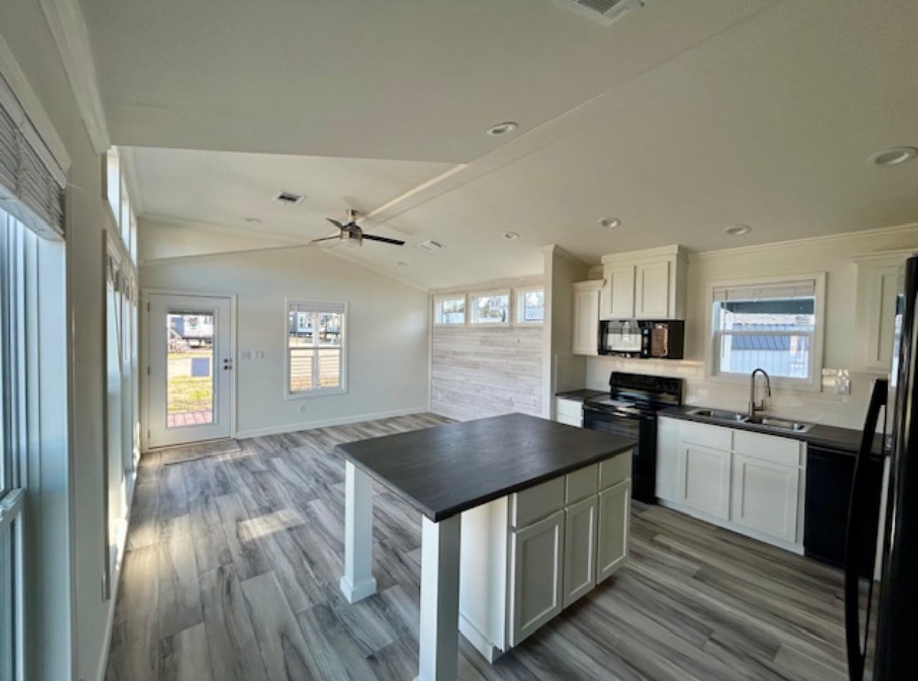 Modern kitchen and living area with vaulted ceiling, central island, wood floors, and ample natural light. White cabinets and black appliances. Cozy and inviting.