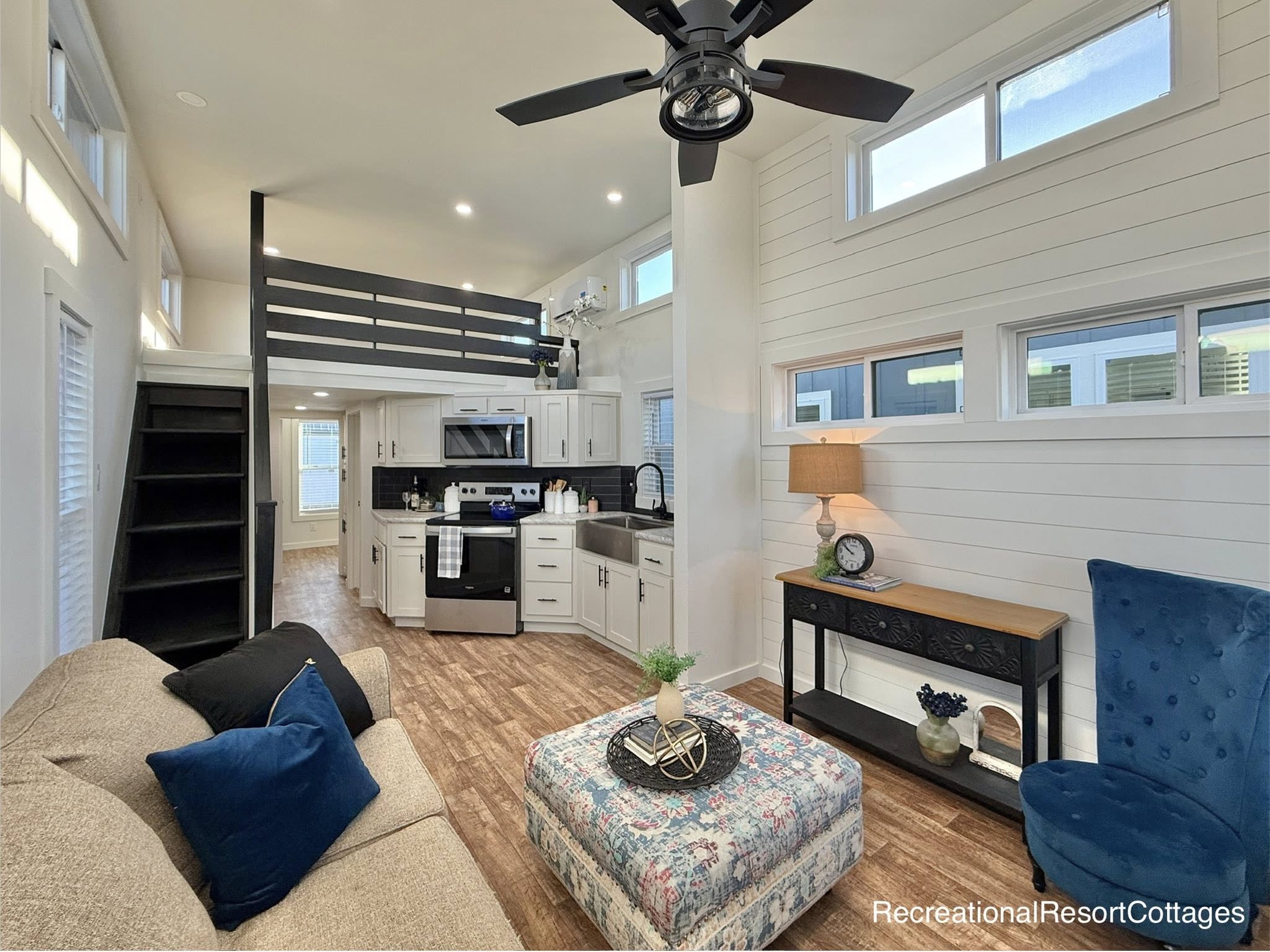 Modern tiny home interior with a cozy style. Features a loft, compact kitchen, plush sofa, blue accent chair, and stylish decor on wooden flooring.