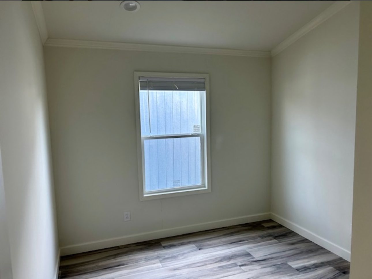 Small empty room with light gray walls, wood-like flooring, and a window with partially open blinds. The space feels clean and minimalist.