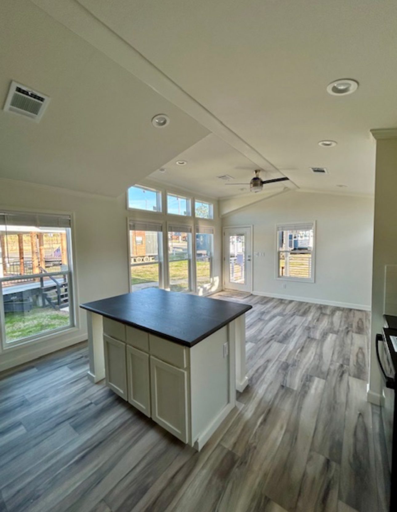 Spacious, modern interior with high ceilings, large windows, and wood-patterned floors. A kitchen island with dark countertop adds elegance. Natural light fills the room.