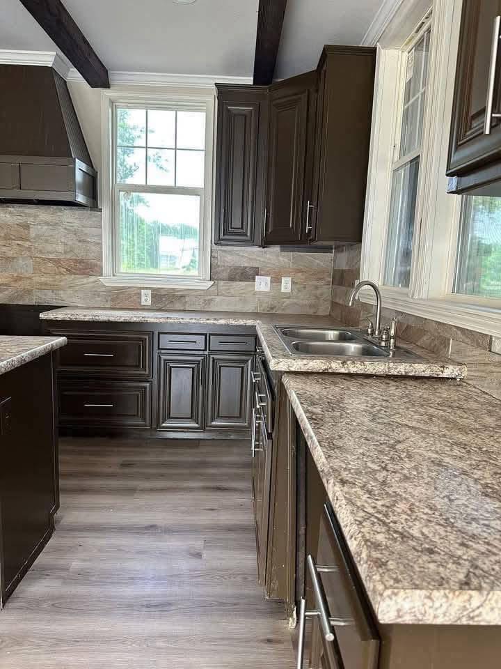 A modern kitchen with dark brown cabinets, light granite countertops, a double sink, and large windows. The light wood floor adds warmth.
