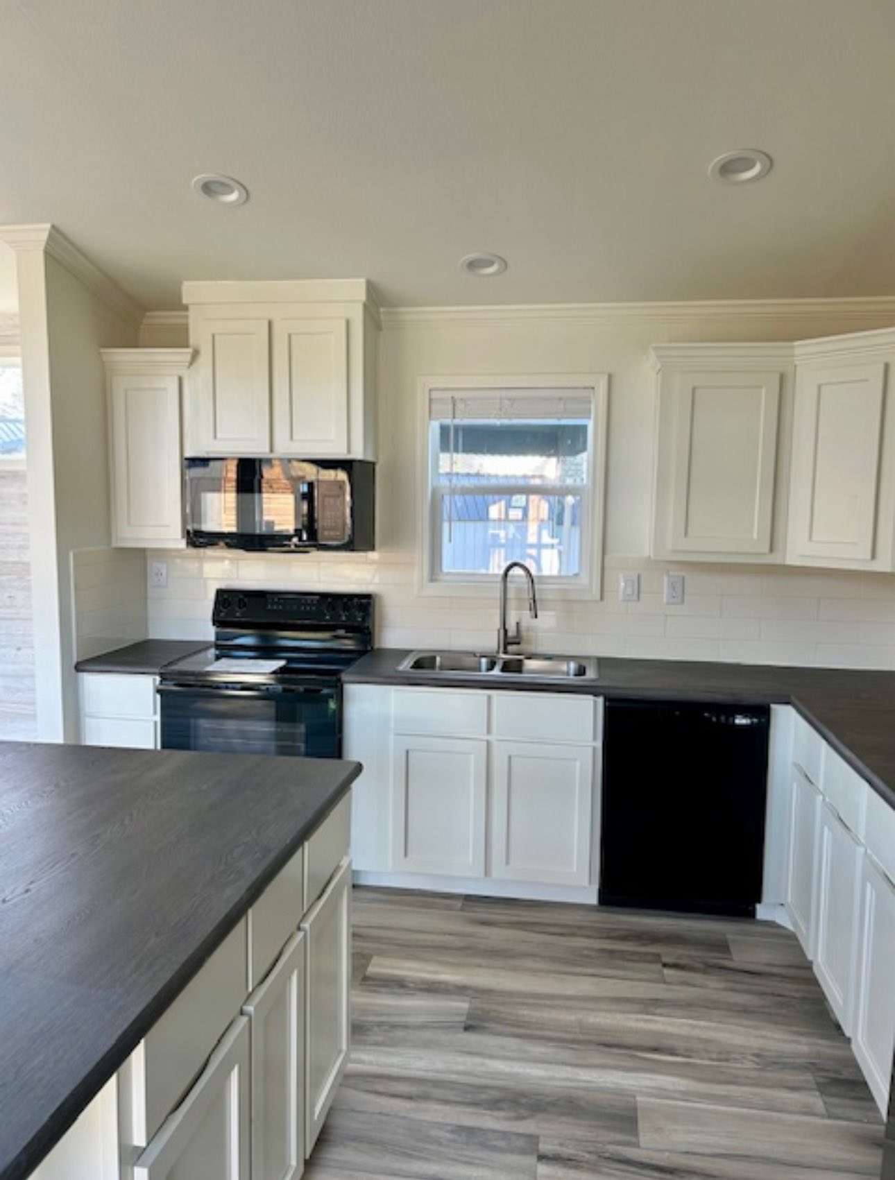 Modern kitchen with white cabinets, dark countertops, and stainless steel appliances. Central island, wood-patterned flooring, and bright lighting.