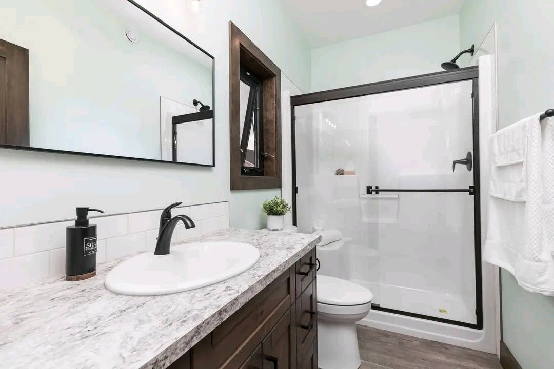 Modern bathroom with a marble countertop, dark wood cabinets, black faucet, large mirror, glass shower door, white tiled walls, and green plant.
