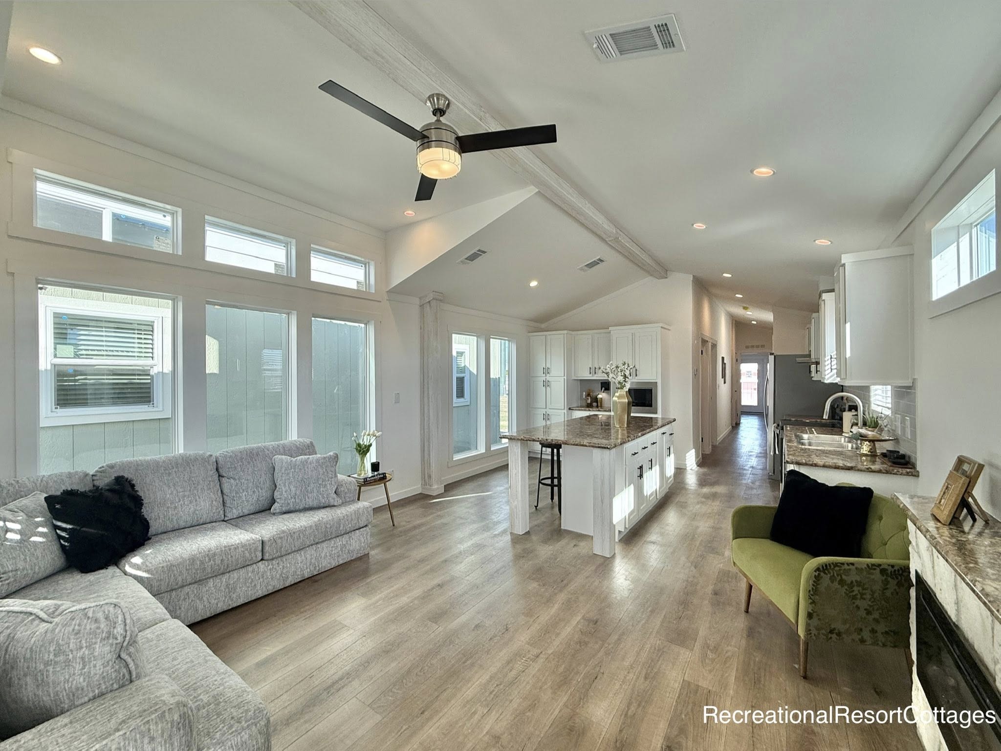 Spacious, modern open-plan living area with light wood floors. Features a gray sofa, green armchair, and kitchen with white cabinets and an island. Abundant natural light and a ceiling fan enhance the airy atmosphere.