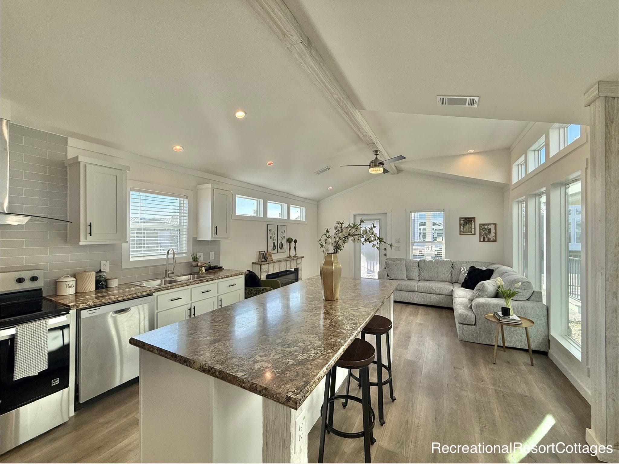 Spacious open-concept living area with large kitchen island, bar stools, a cozy gray sectional, ceiling fan, and bright natural light streaming in.
