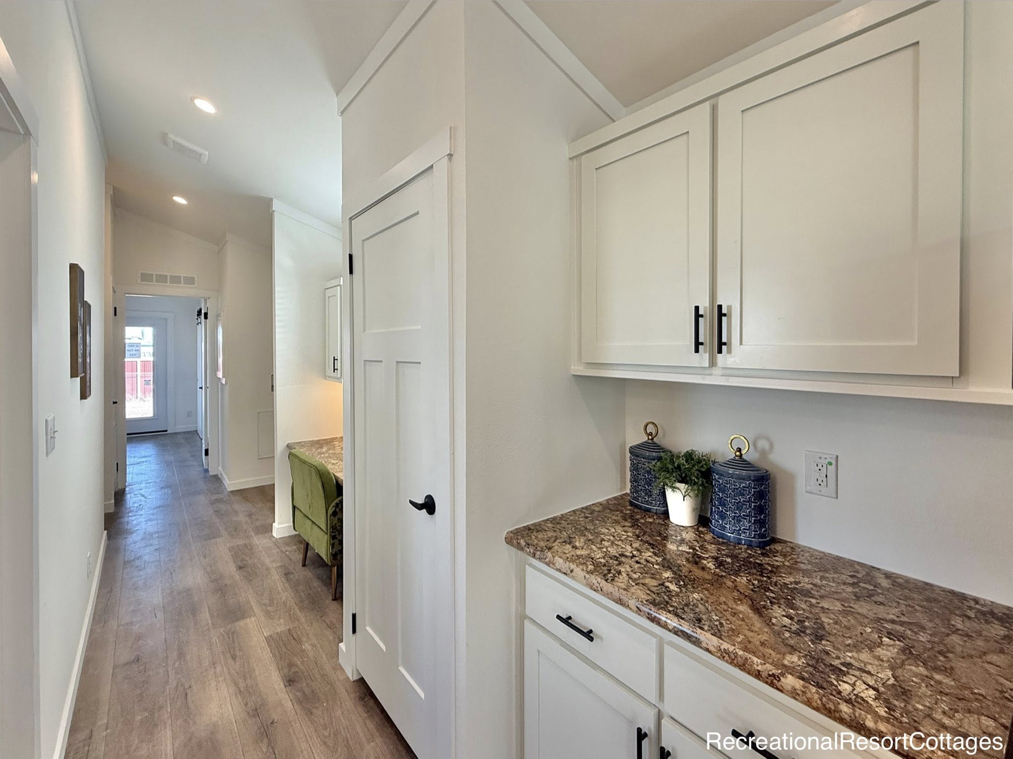 Bright hallway with wood flooring leads to a door with glass panels. White cabinets with brown granite countertops are on the right. Cozy, modern feel.