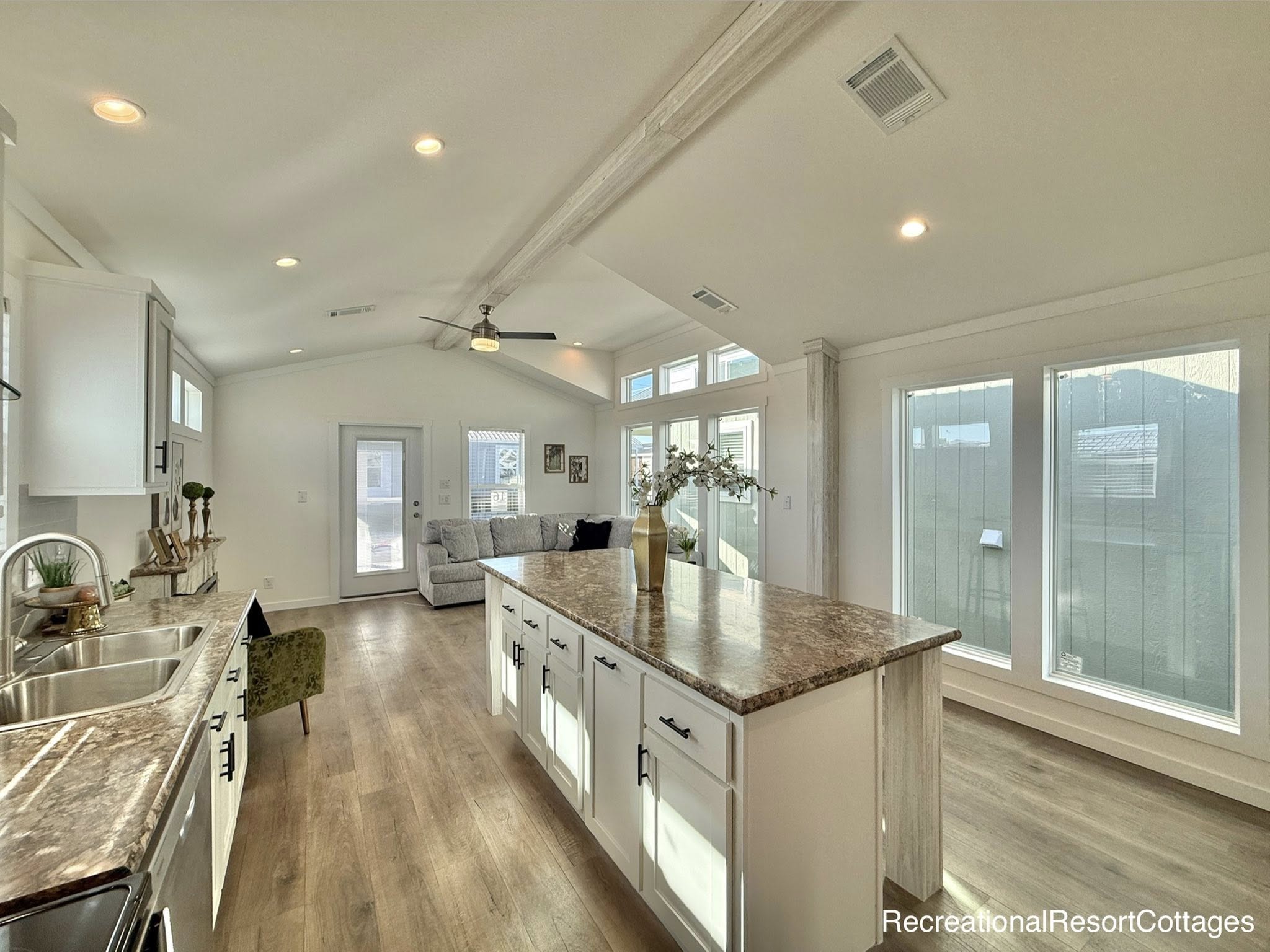 Bright and airy kitchen with white cabinets and a marble island. Sunlight filters through large windows. A cozy living area is visible in the background.