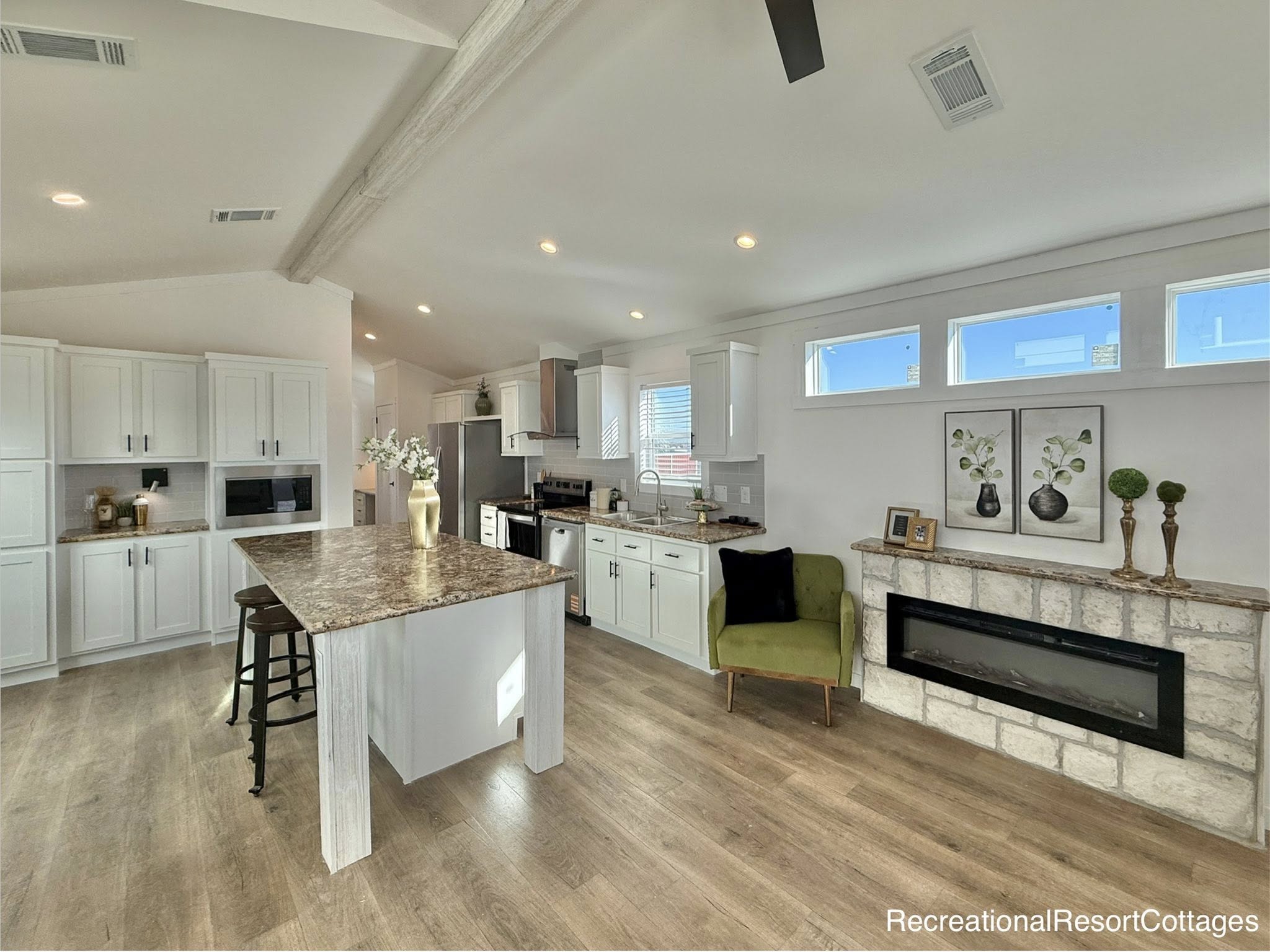 Modern kitchen with a granite island, white cabinets, stainless steel appliances, and wood floors. A green chair and stone fireplace add warmth.