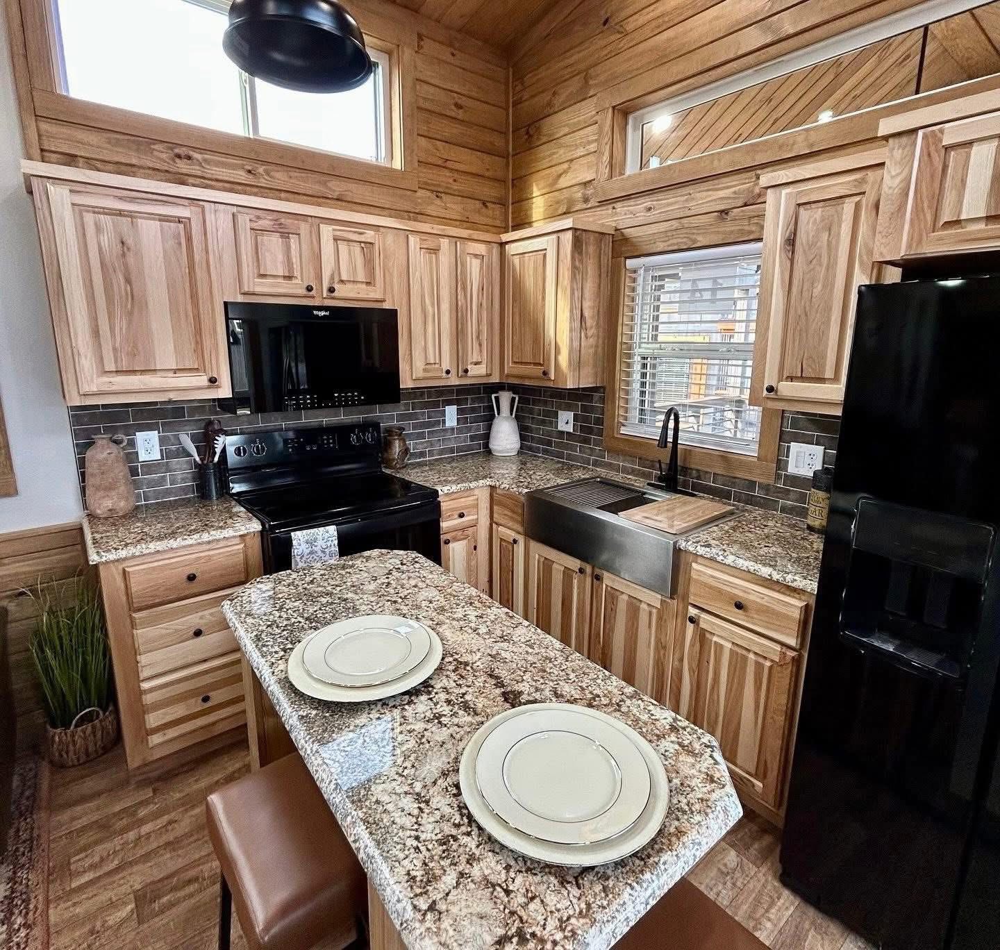 A rustic kitchen with wooden cabinetry, granite countertops, and a black stove and fridge. There is a farmhouse sink and an island with two plates. Sunlight filters through a window, creating a warm, inviting atmosphere.