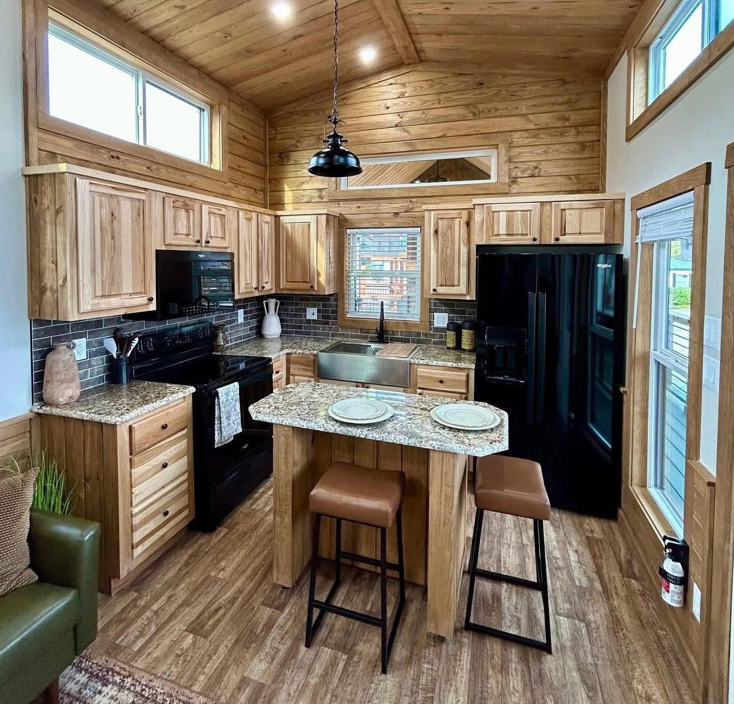 Warm, rustic kitchen with wooden cabinets, a granite island, and a black fridge. Natural light streams in, creating a cozy, inviting atmosphere.