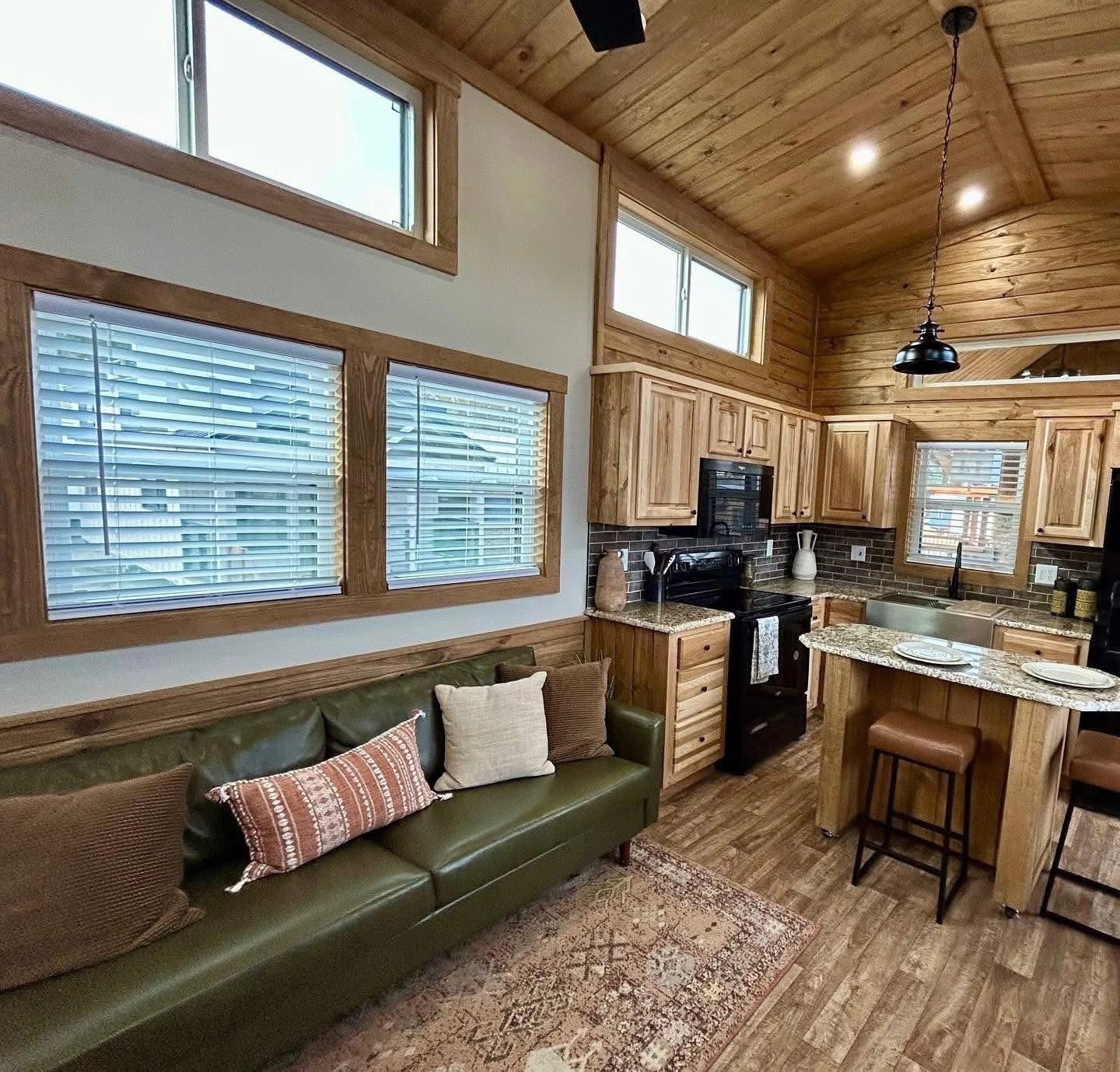 Cozy cabin interior with wood-paneled walls and ceiling, green leather couch with patterned pillows, and kitchen with wooden cabinets and island.