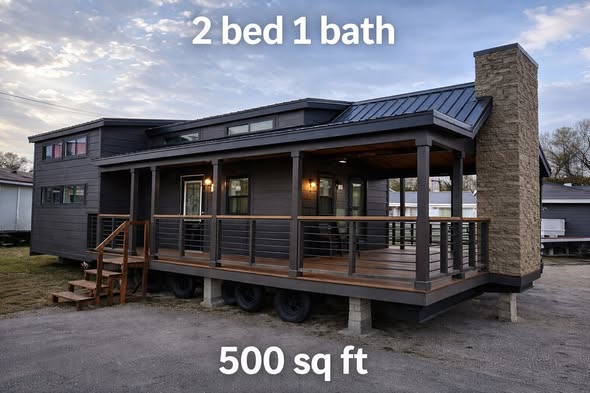 Modern tiny house with dark wood siding, elevated porch, and large stone chimney. Text states: "2 bed 1 bath, 500 sq ft." Cozy and compact design.